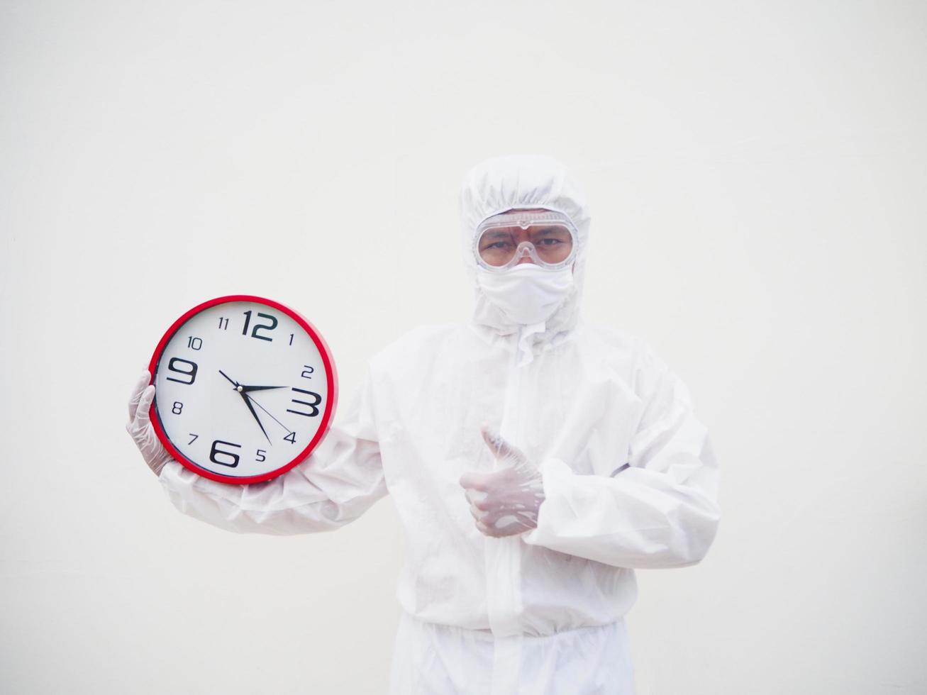 retrato de médico o científico con uniforme de suite ppe sosteniendo un despertador rojo y mirando la cámara con varios gestos. covid-19 concepto aislado fondo blanco foto