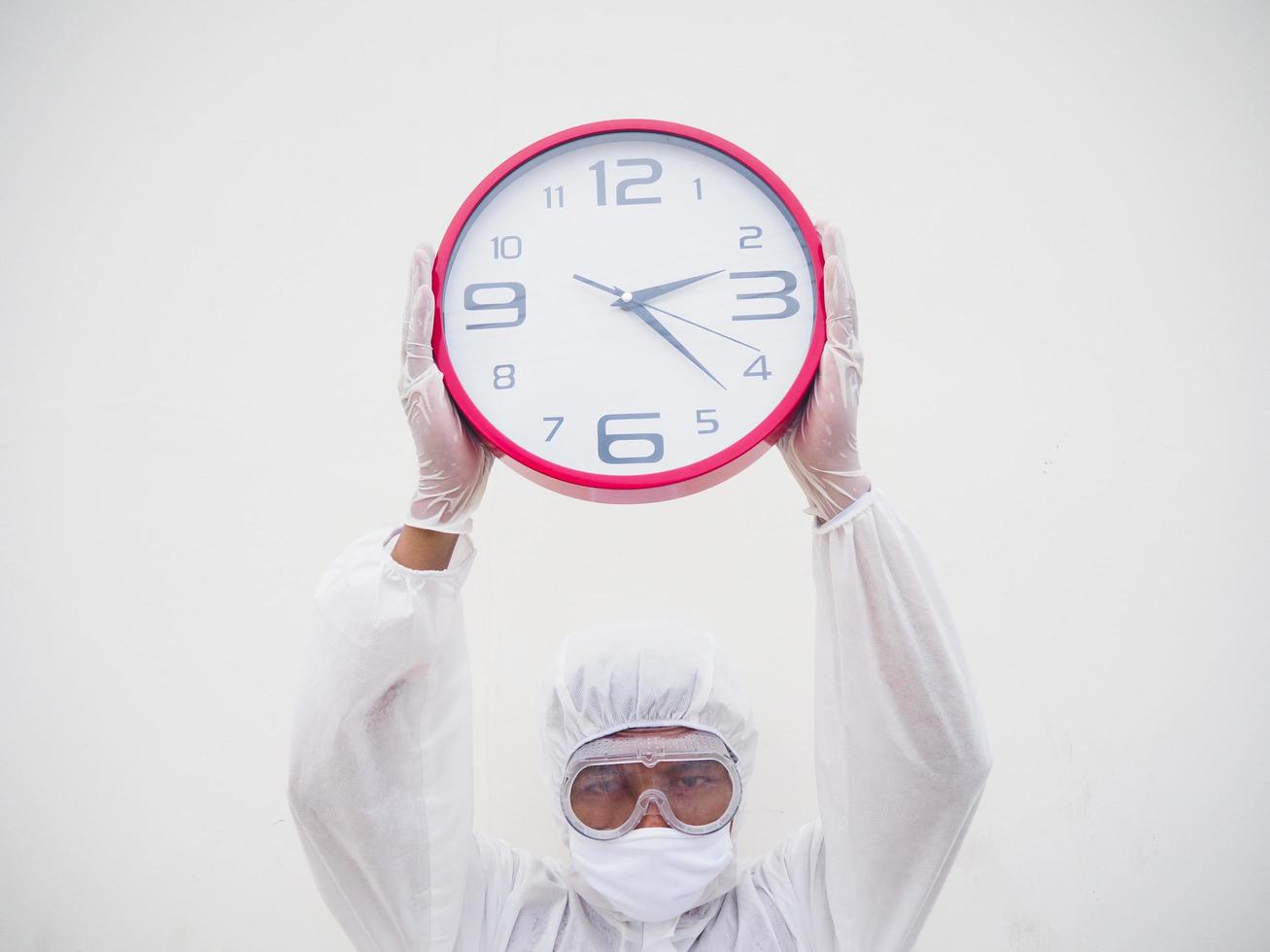 retrato de médico o científico con uniforme de suite ppe sosteniendo un despertador rojo y mirando la cámara con varios gestos. covid-19 concepto aislado fondo blanco foto