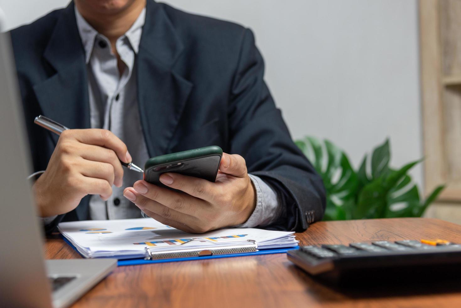 mano de hombre de negocios que sostiene el teléfono móvil y la pluma con gráficos y documentos de negocios en el escritorio. calculadora y computadora para inversión y análisis económico. concepto de negocio financiero y fiscal. foto