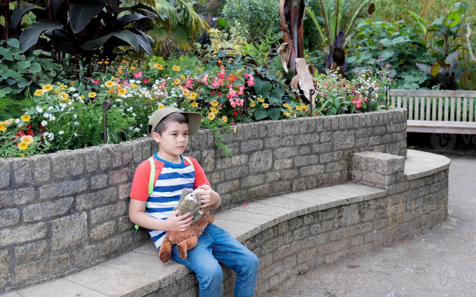 Portrait kid holding plush toy sitting in the garden,Happy boy looking out with thinking face,Child wearing cap hat, carrying backpack enjoying time in the park,Children exploring with nature concept photo