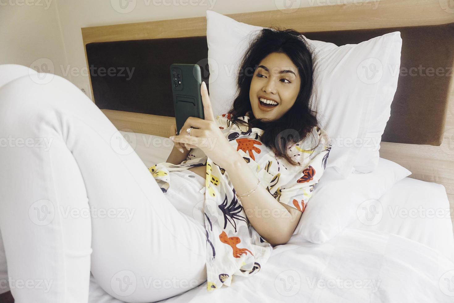 mujer joven feliz revisando su teléfono mientras está acostada en la cama foto