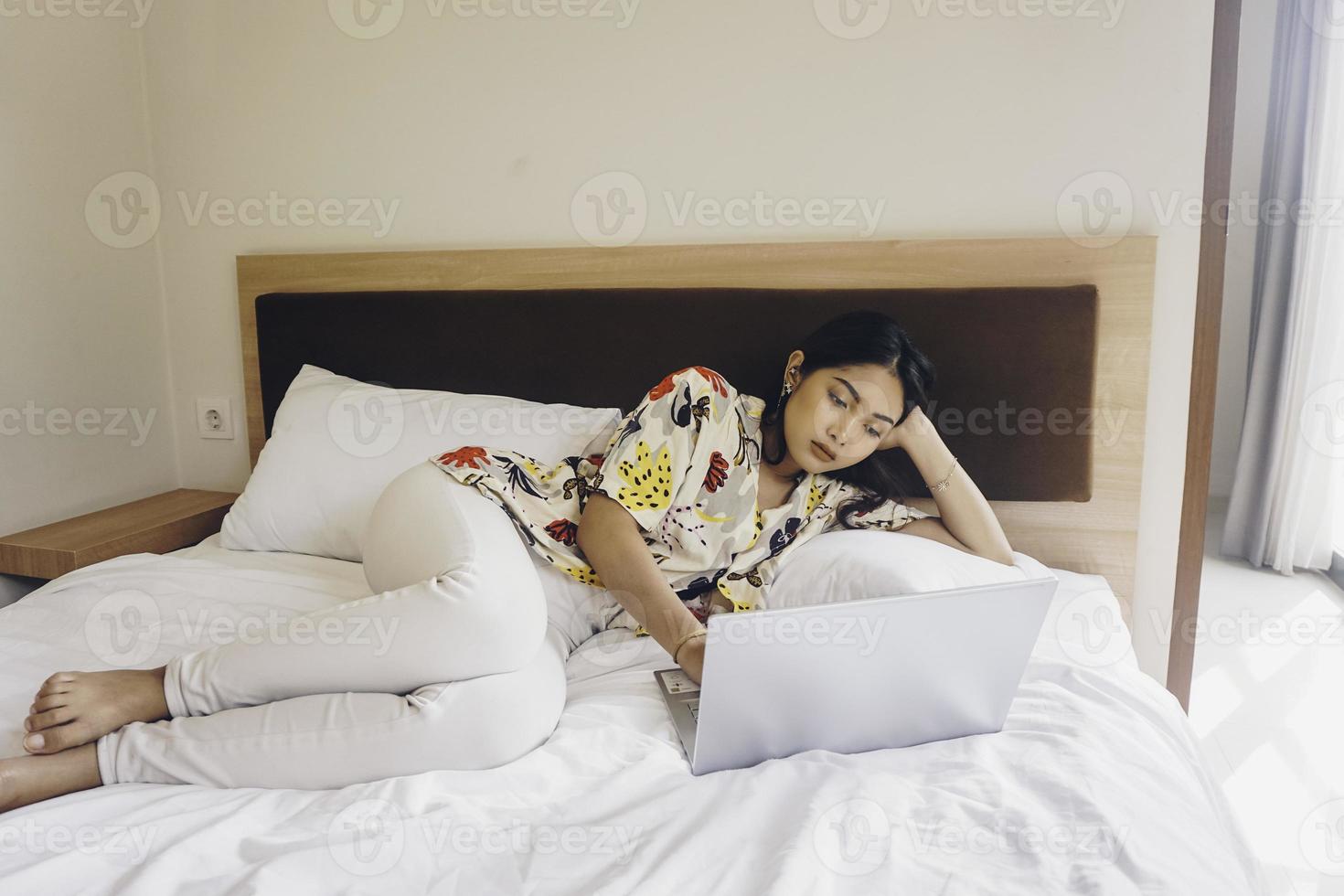 Bored young woman working from the bed. Hold the laptop on the knees and type on the keyboard. photo