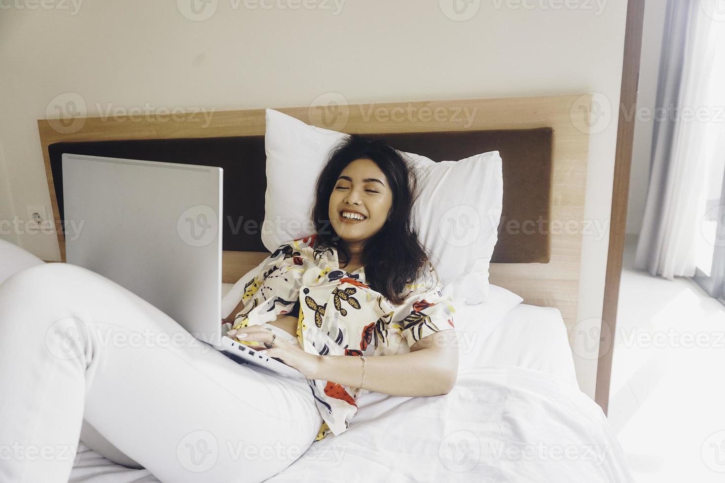 Happy young woman working from the bed. Hold the laptop on the knees and type on the keyboard. photo