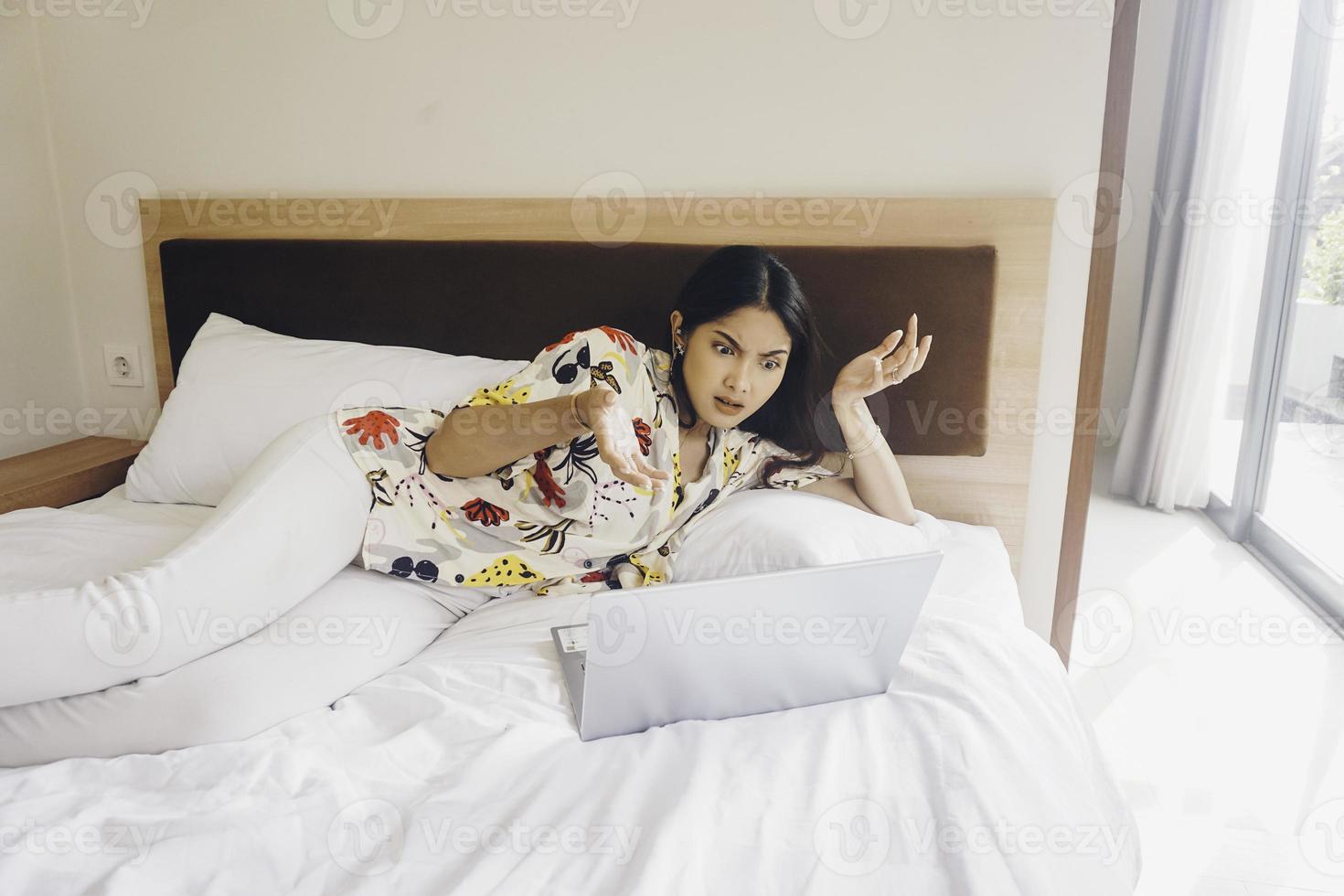 Upset young woman working from the bed. Hold the laptop on the knees and type on the keyboard. photo