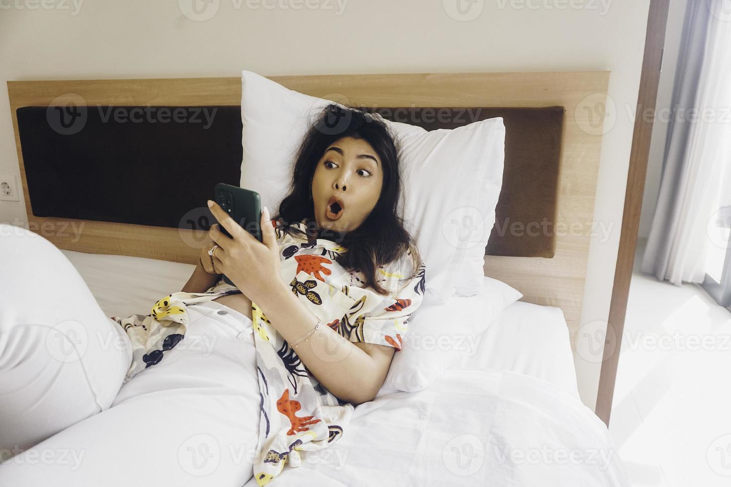 mujer joven sorprendida revisando su teléfono mientras está acostada en la cama foto