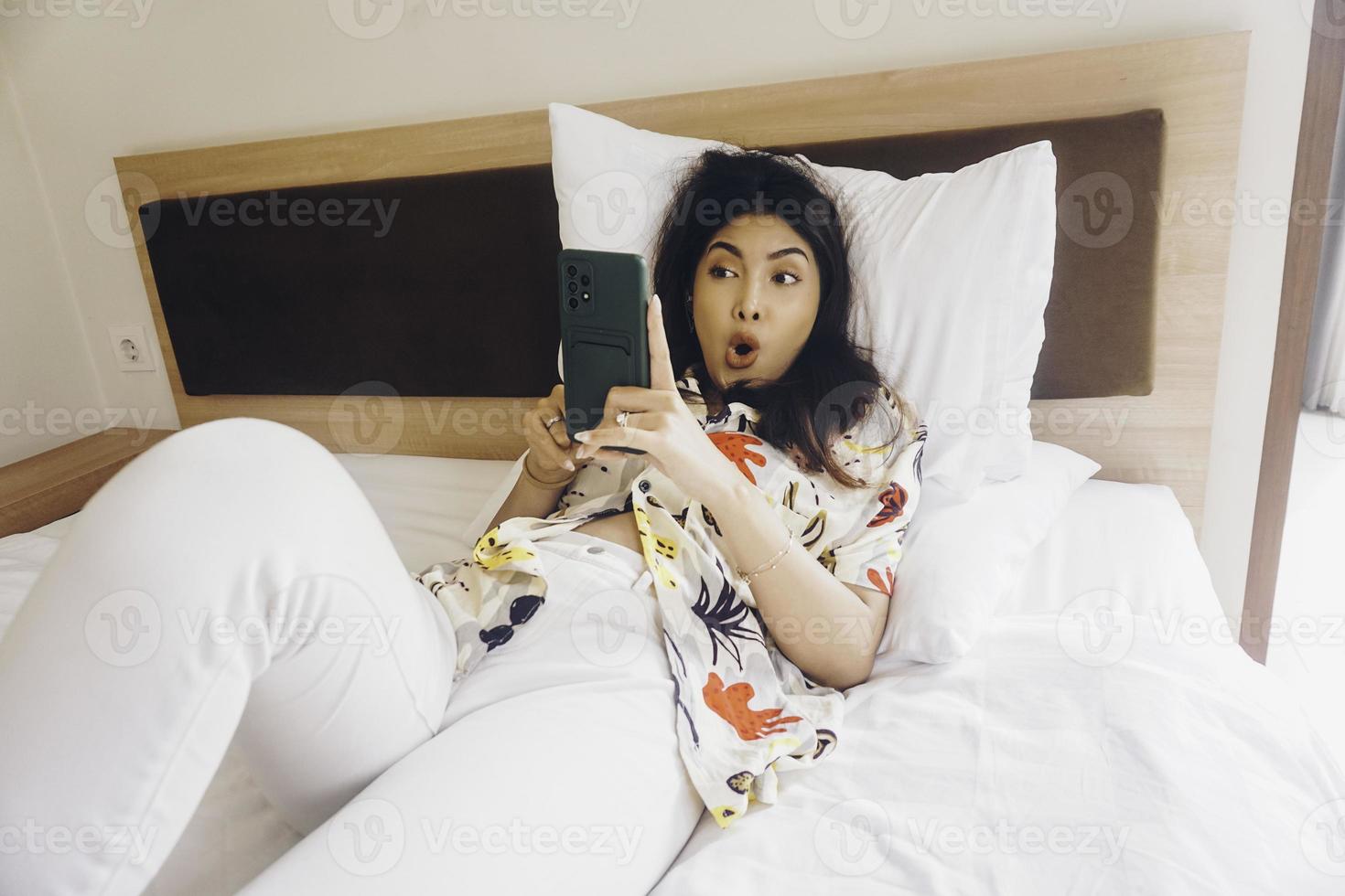mujer joven sorprendida revisando su teléfono mientras está acostada en la cama foto