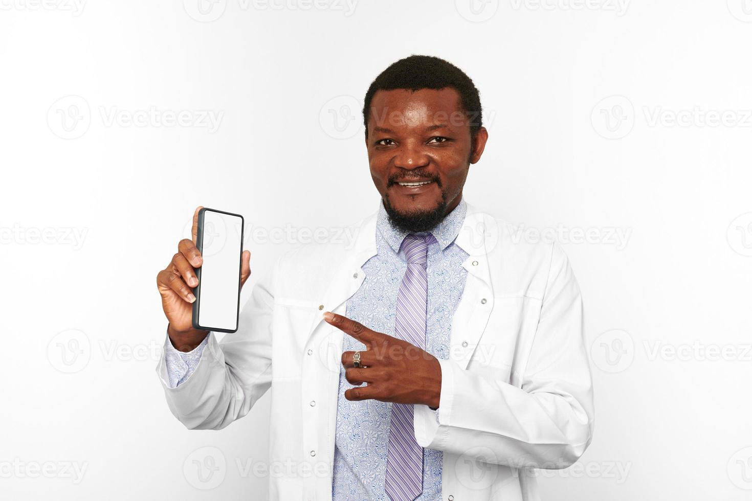 Smiling black doctor man with small beard in white coat holds smartphone blank mockup photo