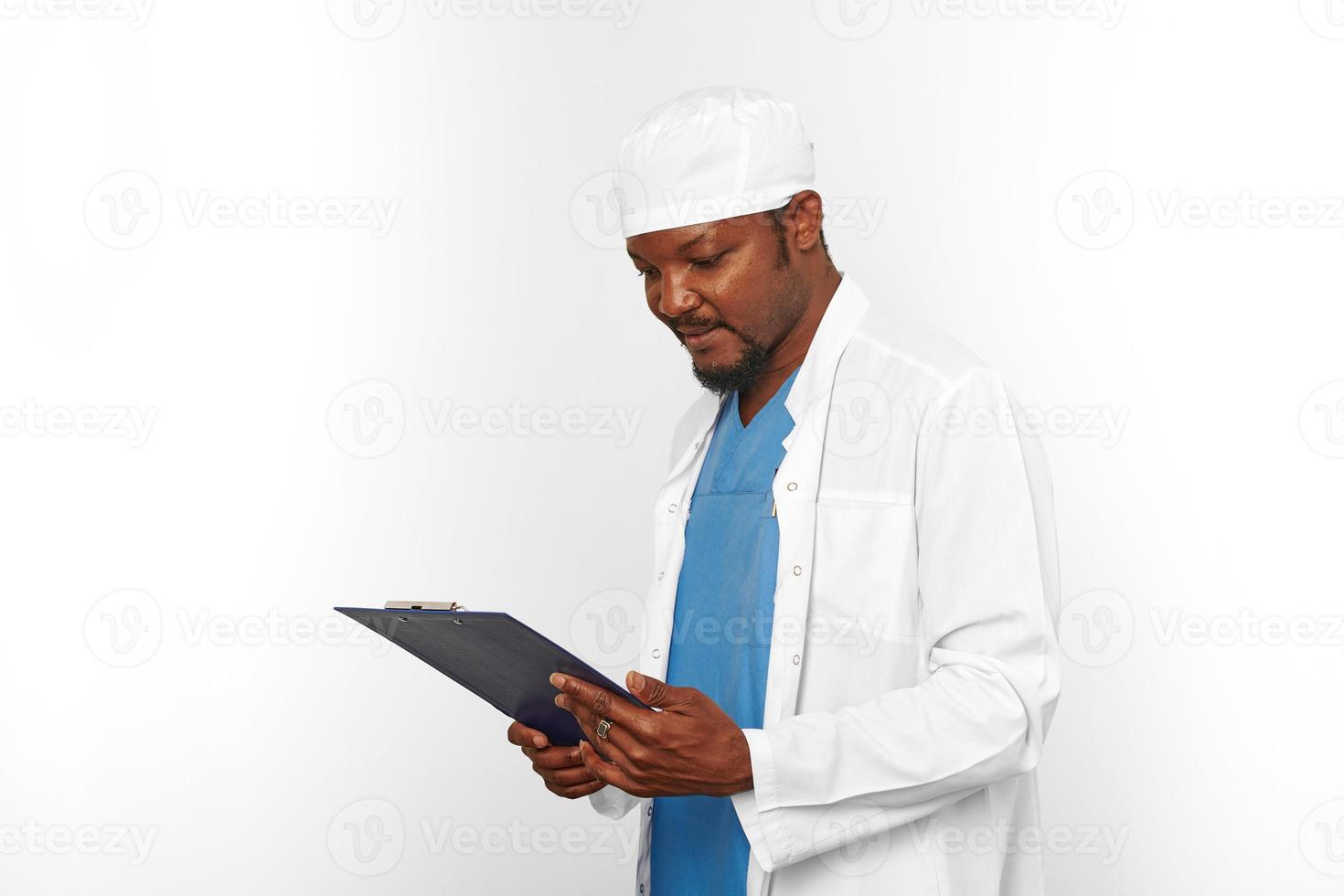 Black surgeon doctor bearded man in white coat and cap looks to medical chart on clipboard on white photo