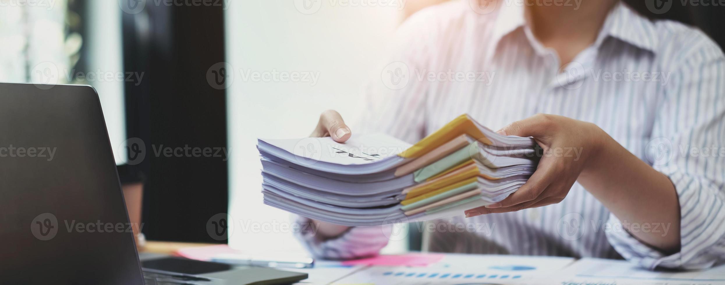Business Documents concept  Employee woman hands working in Stacks paper files for searching and checking unfinished document achieves on folders papers at busy work desk office. Soft focus photo