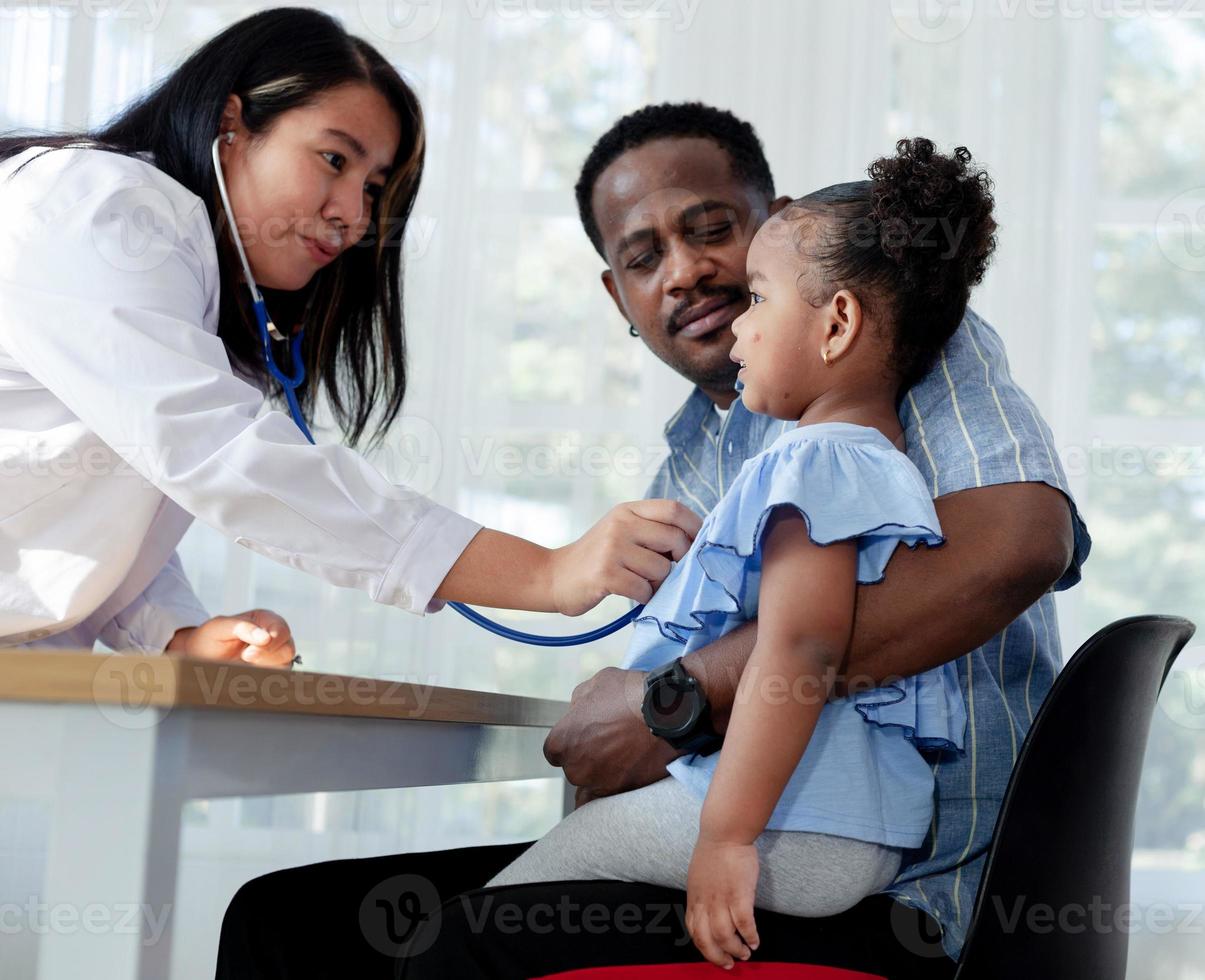 un padre y una hija afroamericanos hablan con una doctora que examina el estado de una niña enferma y linda. centrarse en los niños foto