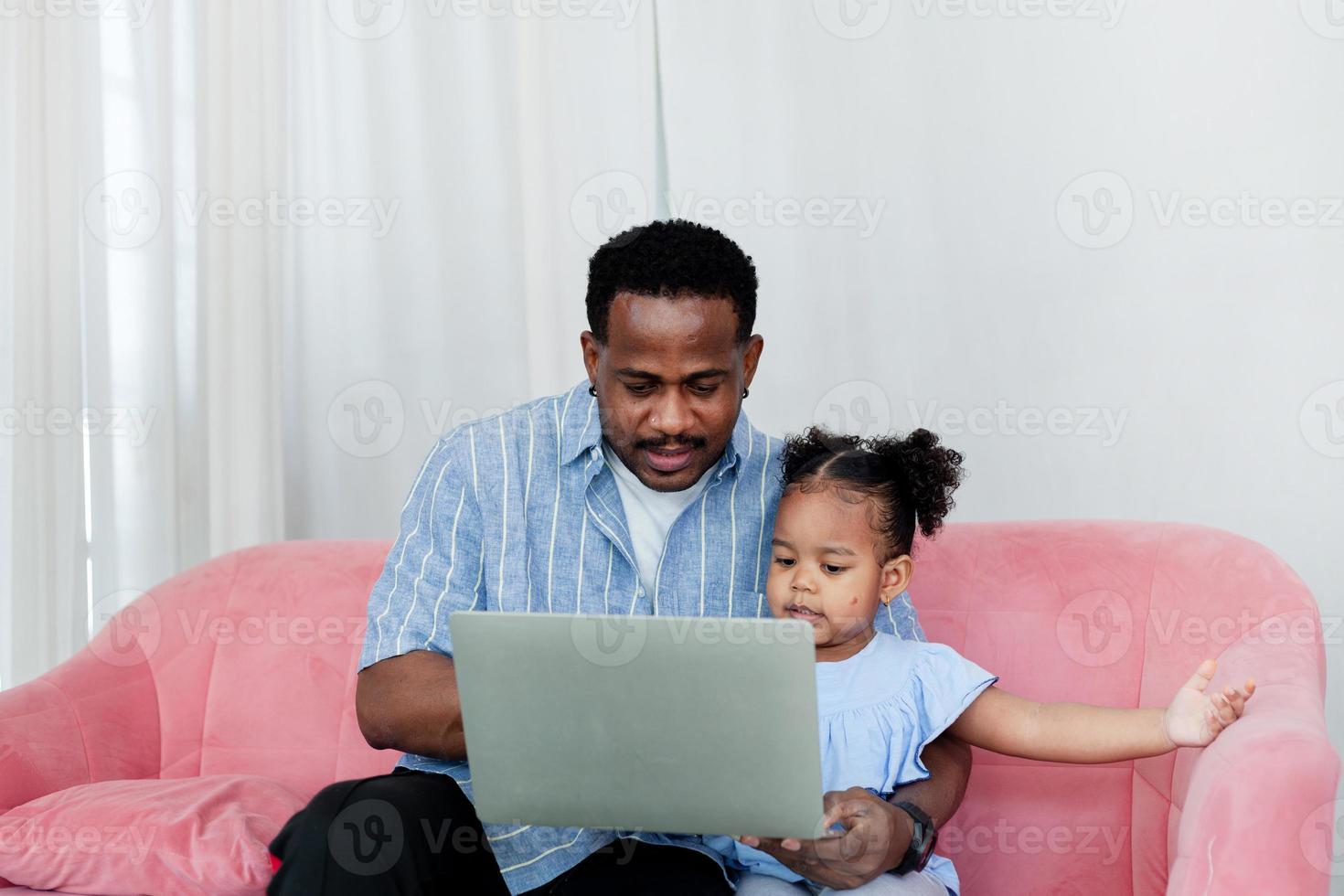 Happy african father and little daughter wearing a blue shirt using laptop doing online shopping on sofa at home . Internet technology concept. photo