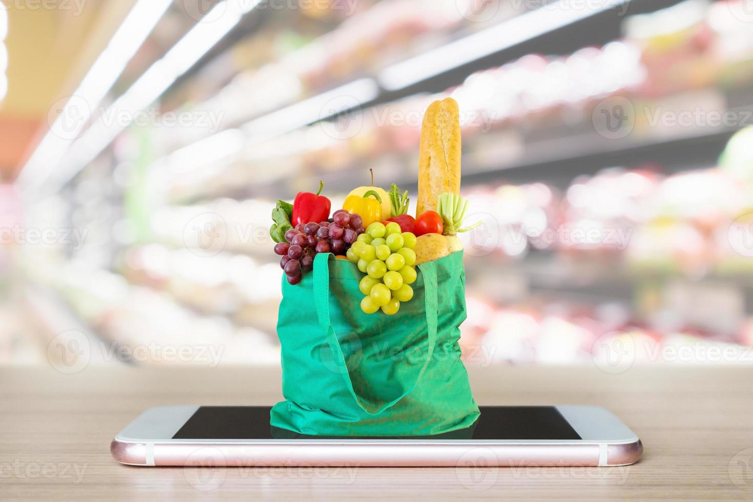 comida fresca y verduras en una bolsa de compras verde en un teléfono inteligente móvil en una mesa de madera con pasillo de supermercado fondo borroso concepto en línea de comestibles foto