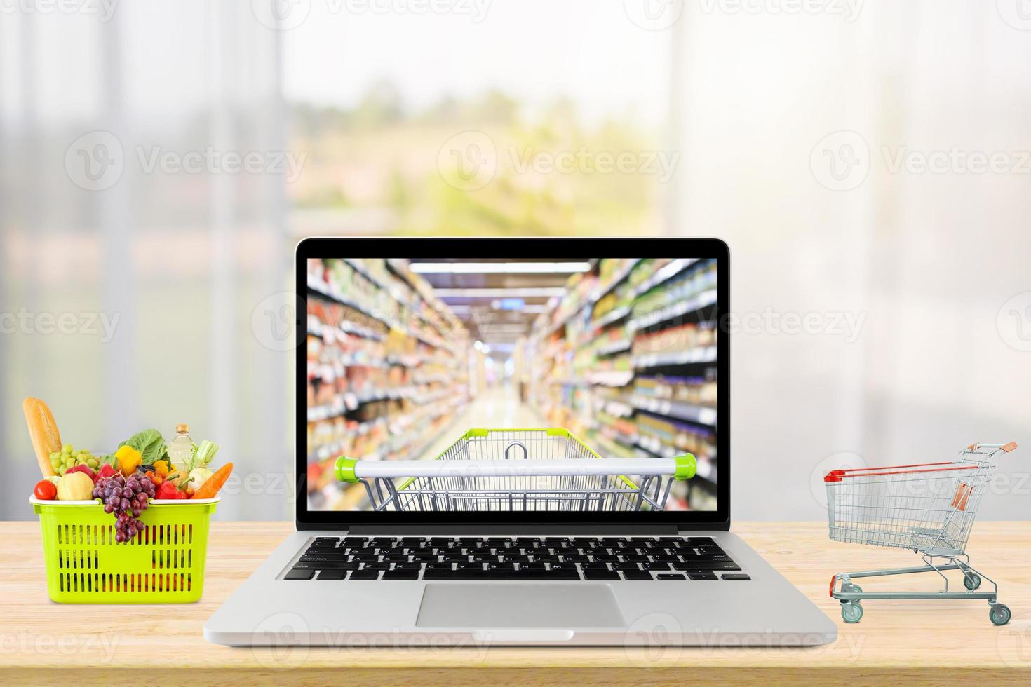 laptop computer and shopping cart on wood table with window curtain abstract blur background grocery online concept photo