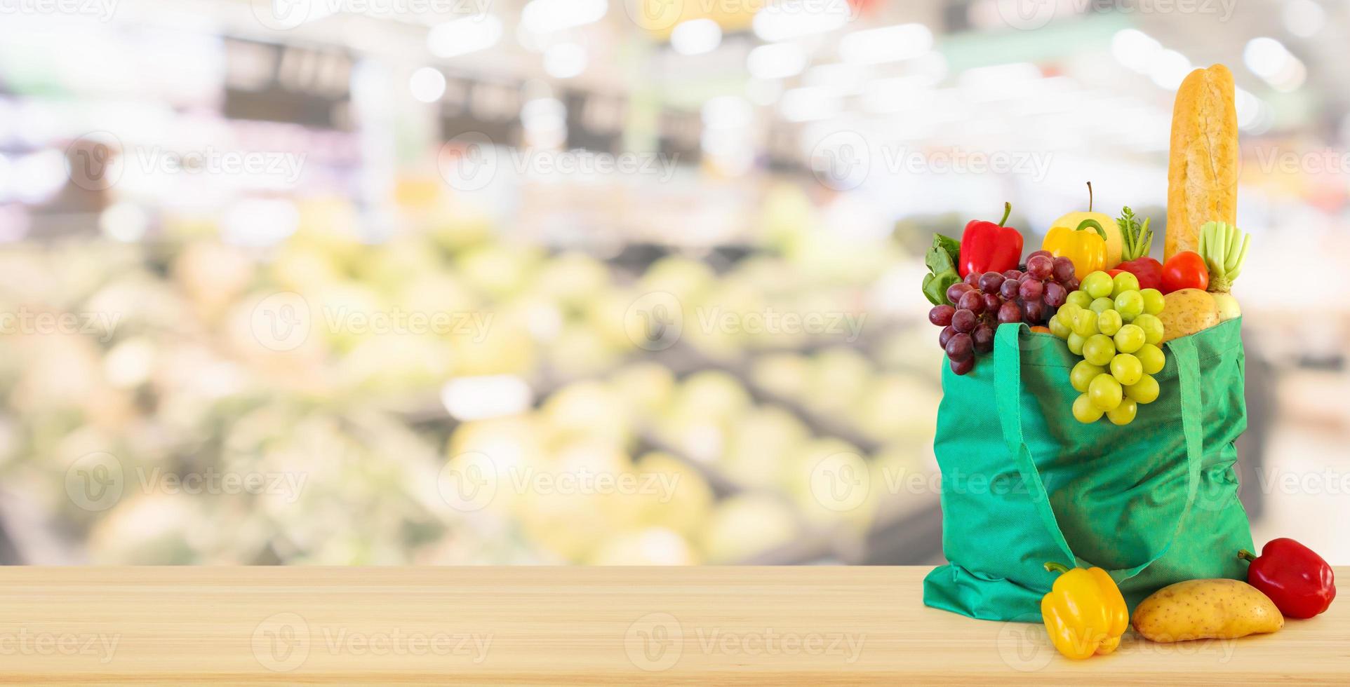 Fresh fruits and vegetables in reusable green shopping bag on wood table top with supermarket grocery store blurred defocused background with bokeh light photo
