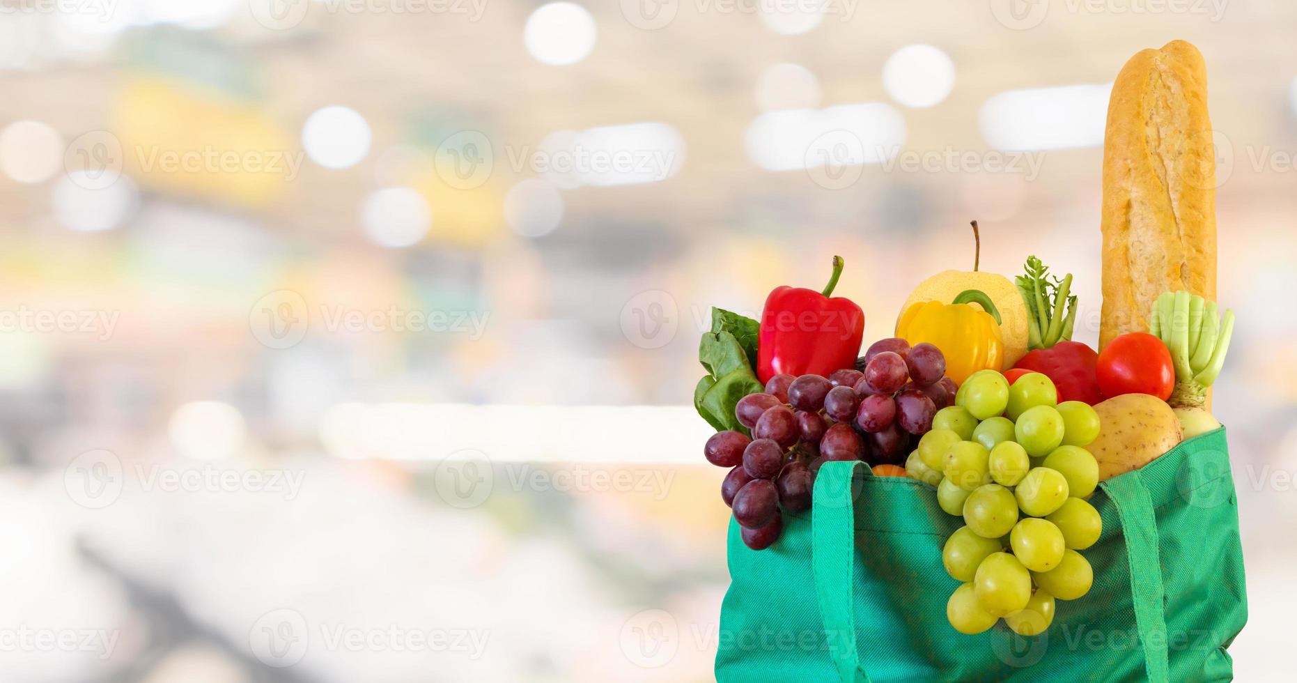 Fresh fruits and vegetables in reusable green shopping bag with supermarket grocery store blurred defocused background with bokeh light photo