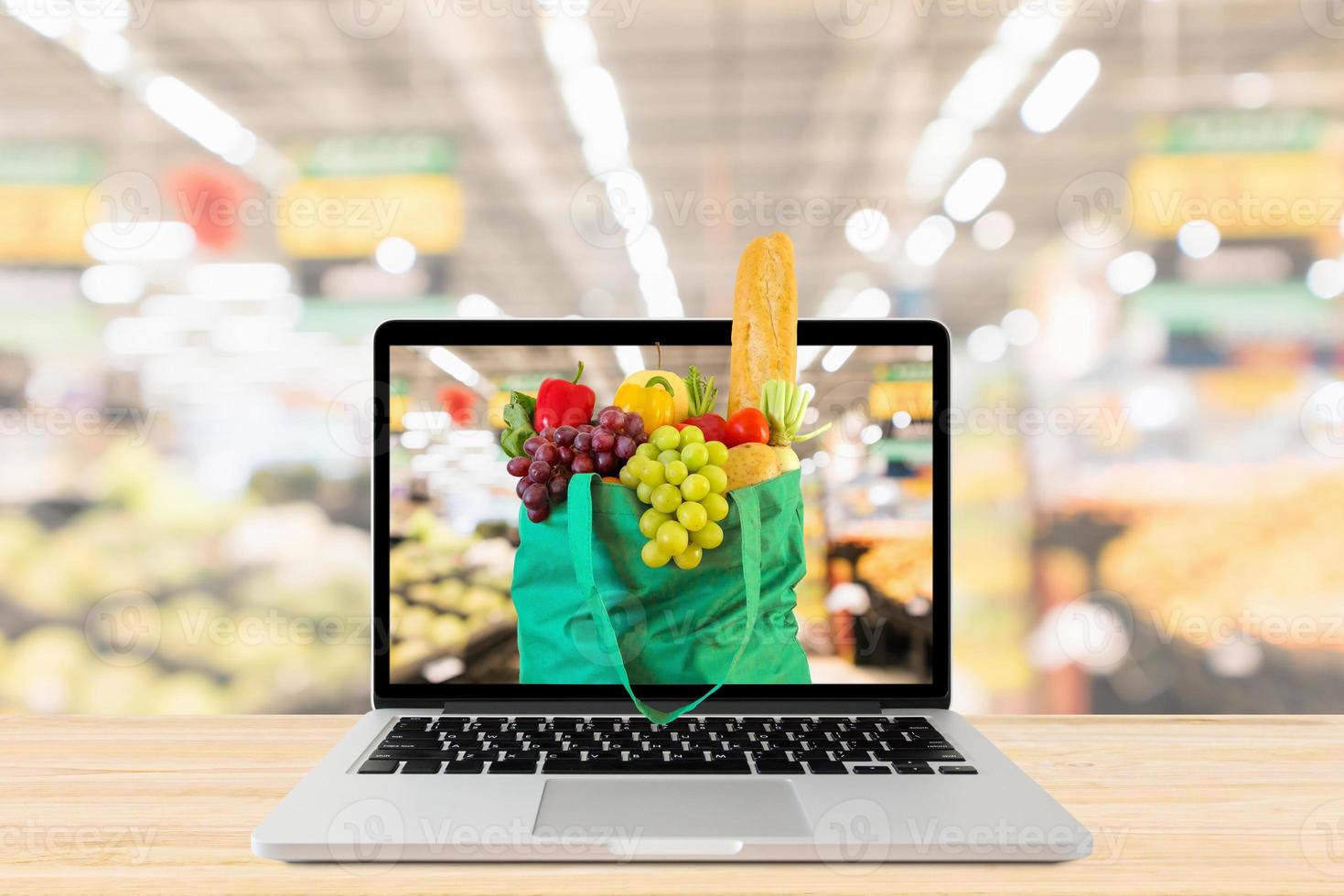 pasillo de supermercado fondo borroso con computadora portátil y bolsa de compras verde en mesa de madera concepto en línea de comestibles foto