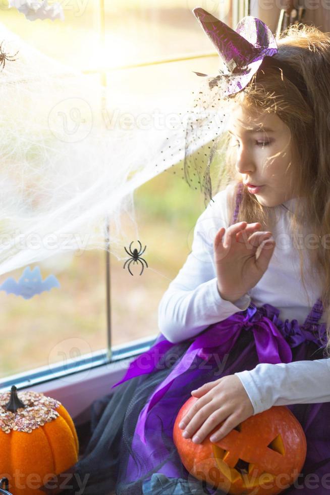 una niña disfrazada de bruja en el alféizar de la ventana está jugando con una telaraña y una araña, decorando la casa para la fiesta de halloween. terrible paisaje, miedo y horror, calabazas foto