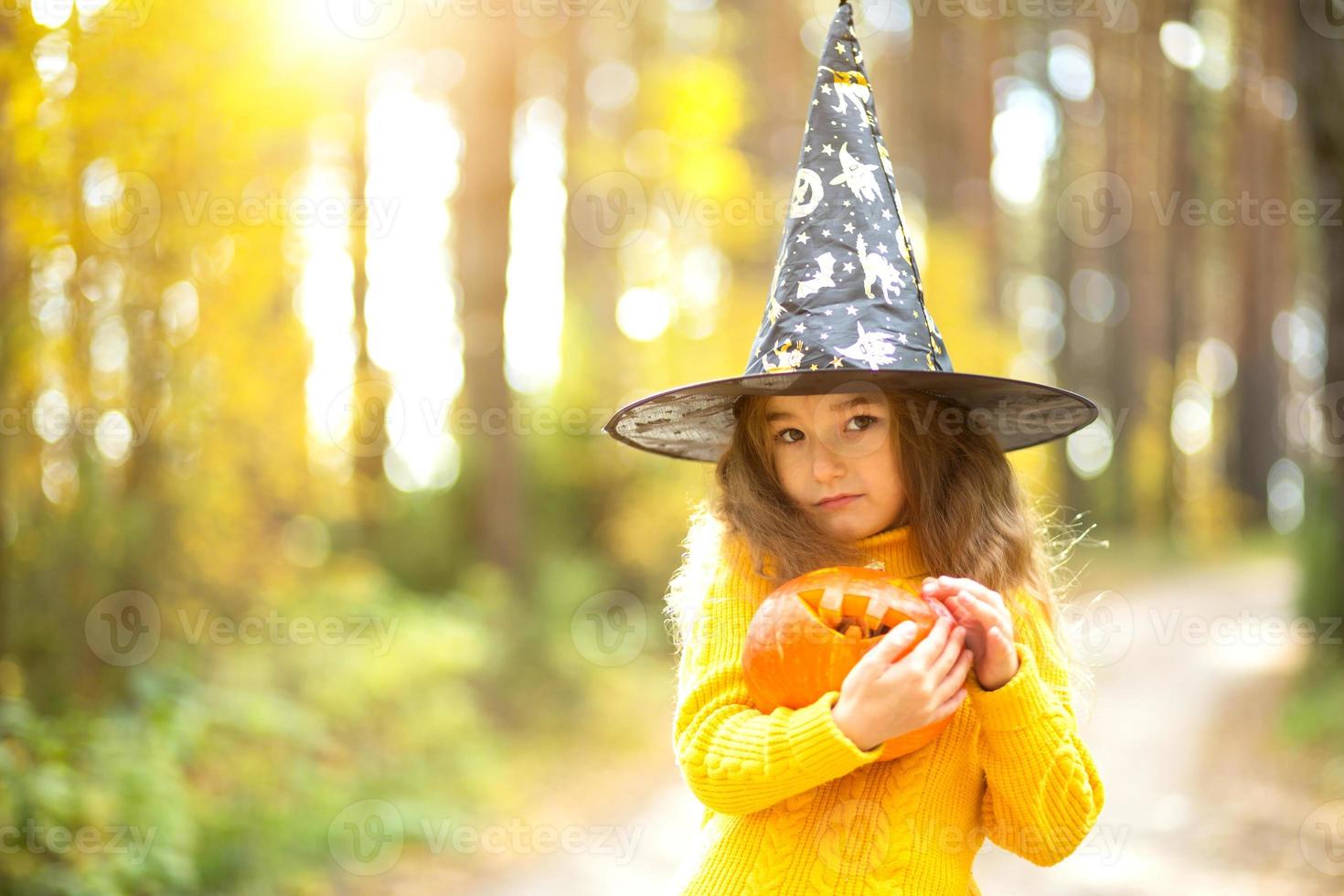 A girl in a witch's hat, in a yellow sweater and with a pumpkin with carved eyes and a mouth Jack o Lantern in a yellow autumn forest. Halloween Holiday photo