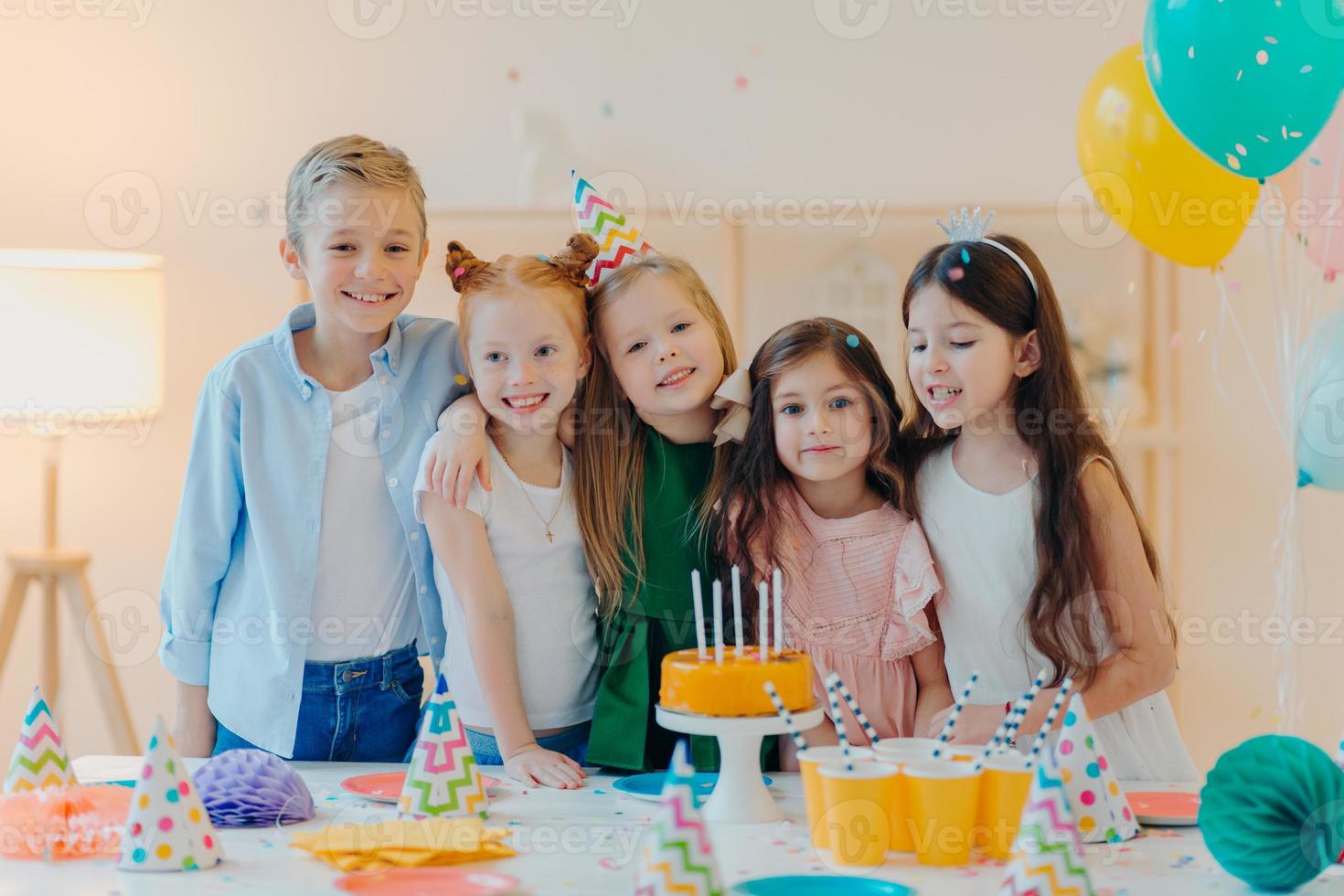 Horizontal shot of group of little children gather together to celebrate birthday, embrace and pose at camera, prepare for special occasion, stand near table with cake, paper cups, party caps photo