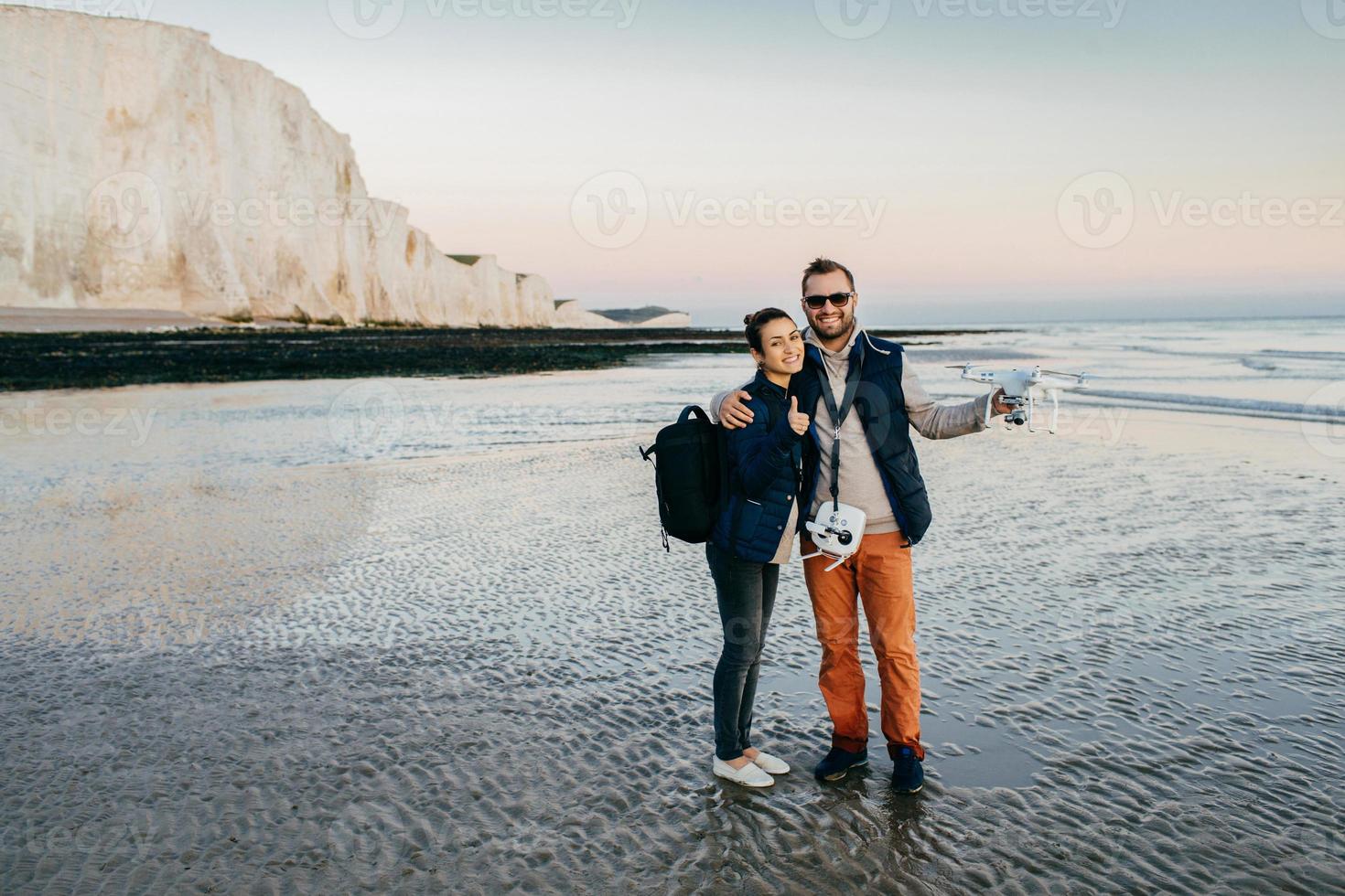 Happy couple hold camera drone air quadrocopter, spend time on seaside, make photo and video of beautiful marine or sea landsacpes. Young man with professionally photography drone outdoors