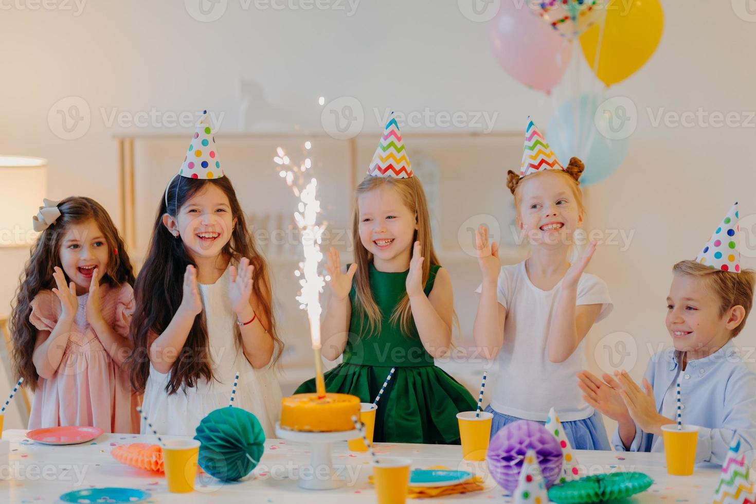 Friends on birthday party, look gladfully at cake, stand near festive table, wear cone hats, clap hands, play together, pose in decorated room. Childhood and holiday concept photo