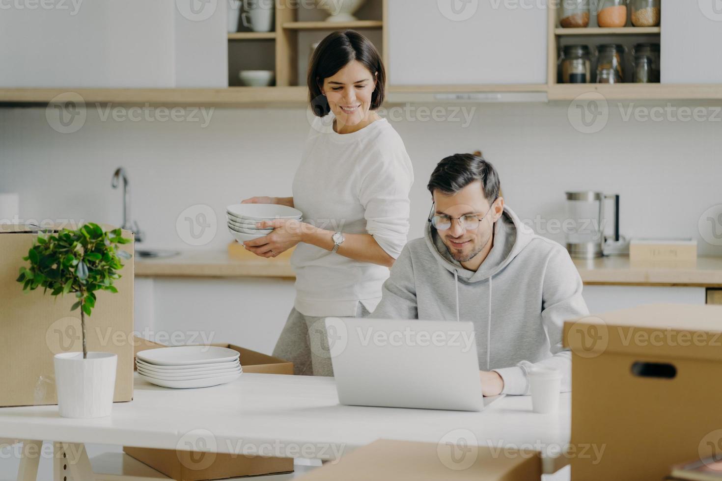 Young brunette woman unpacks boxes with personal belongings, places plates in cupboard, looks attentively at laptop computer where husband works. Family couple relocate in new modern apartment photo