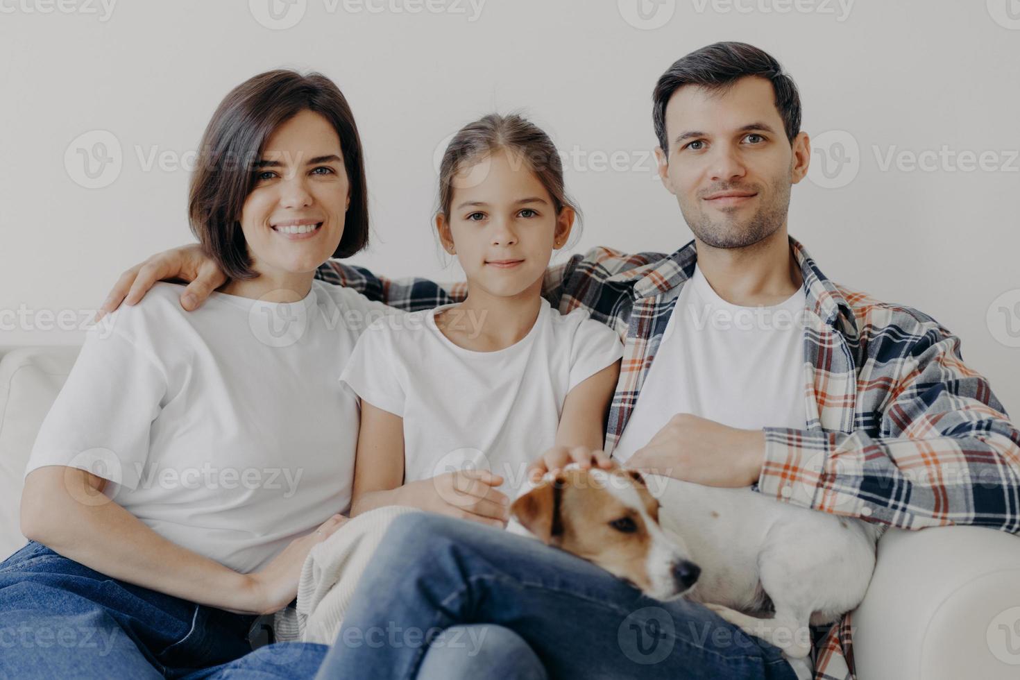 Portrait of pleasant looking mother, dad and small child, sleeping pet, pose all together at sofa against white wall, dressed in casual clothes, spend weekend at home, have friendly relationship photo