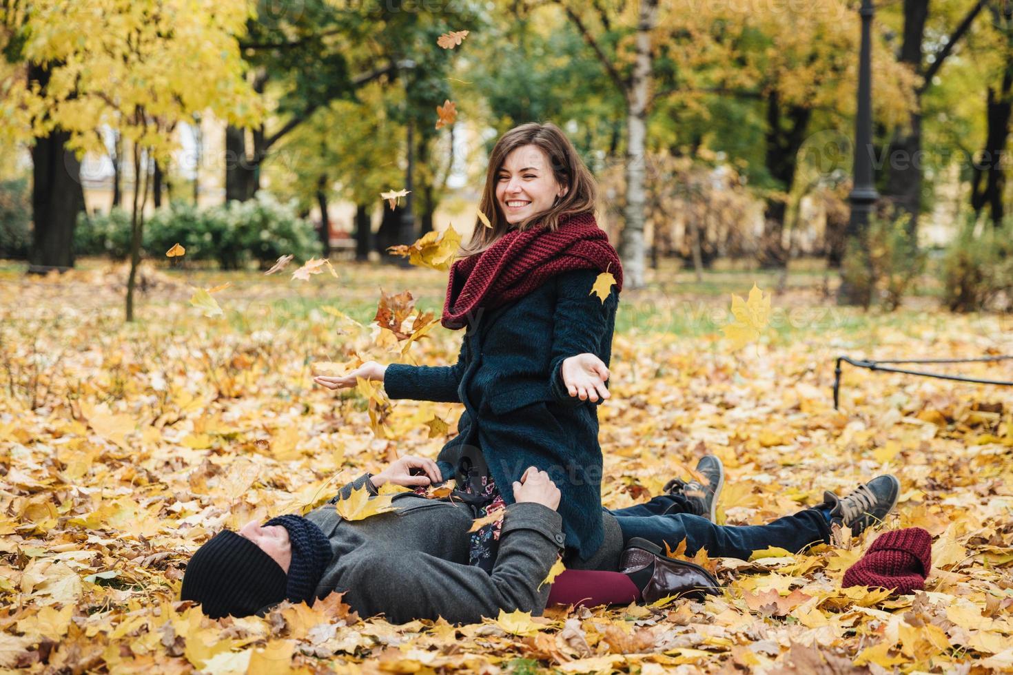 Positive woman wears knitted scarf, sits on boyfriend, throws yellow leaves in air, has good mood as enjoys last warm autumn days. Romantic couple spend weekends together, have fun. Love concept photo
