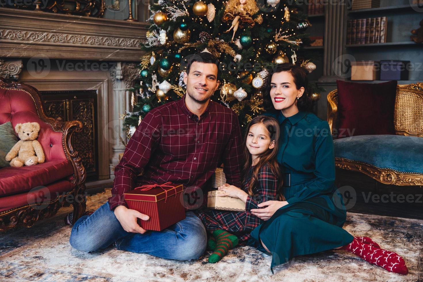 Indoor shot of young family have good relationship, sit together on floor near decorated New Year tree, hold present, congratulate each other with coming New Year and Christmas. Holiday concept photo