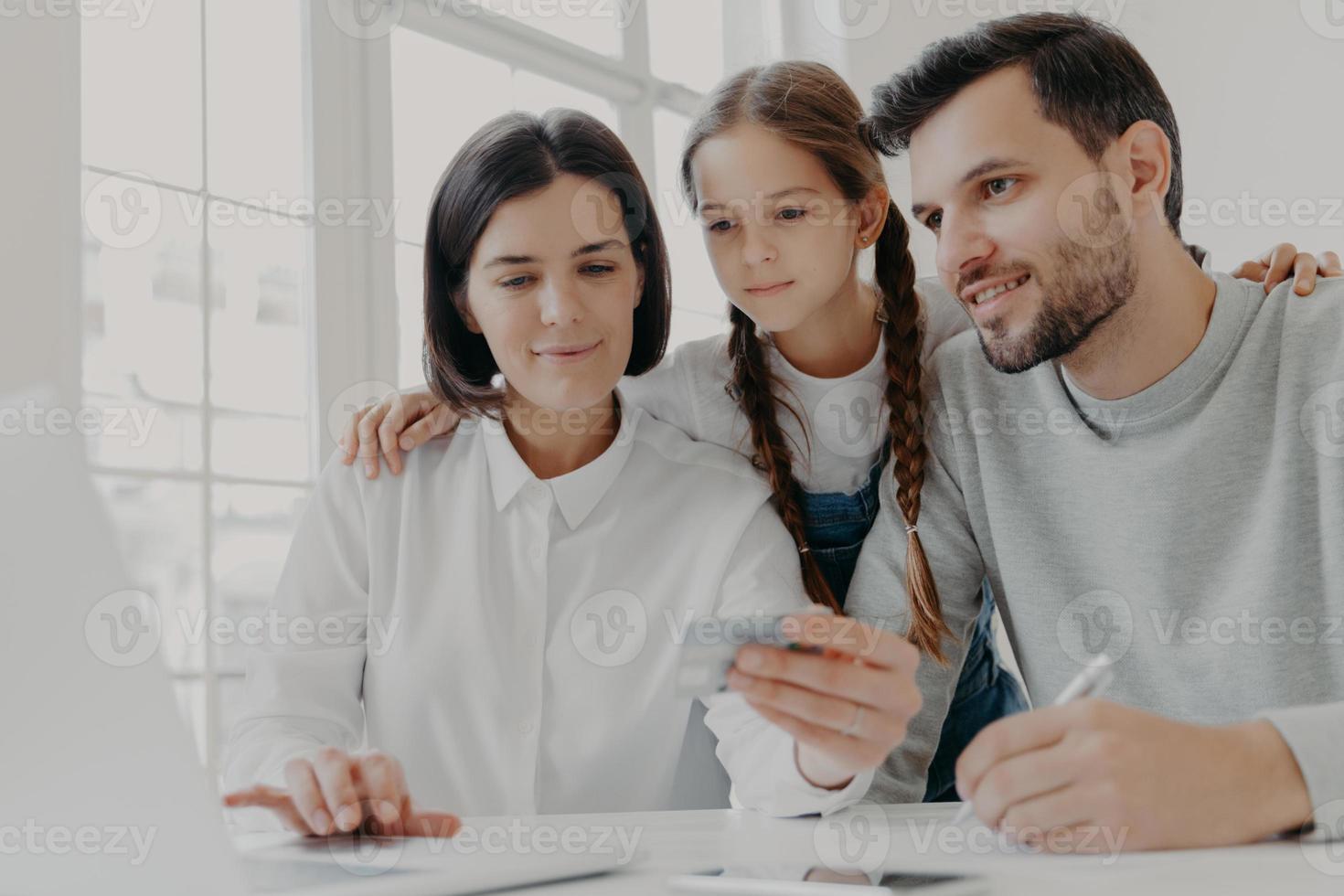 Happy friendly family of father, mother and daughter sit in front of computer, check balance on credit card, make shopping online, buy something necessary, use high speed internet connection. photo