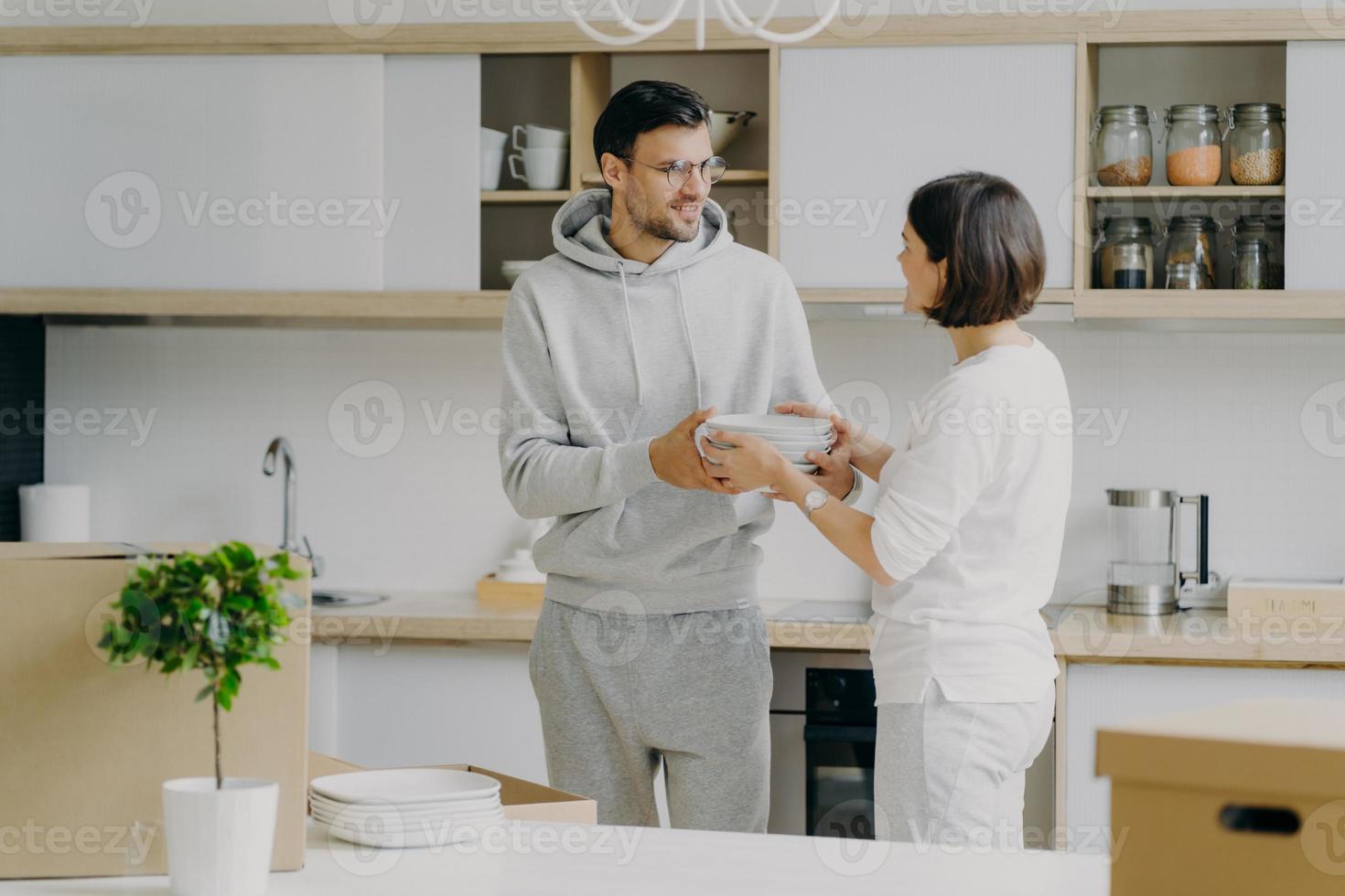 foto de marido y mujer felices desempacando cosas después de mudarse a una nueva casa, llevar platos blancos, posar contra el interior de la cocina, divertirse, vestidos con ropa doméstica. personas y concepto de movimiento