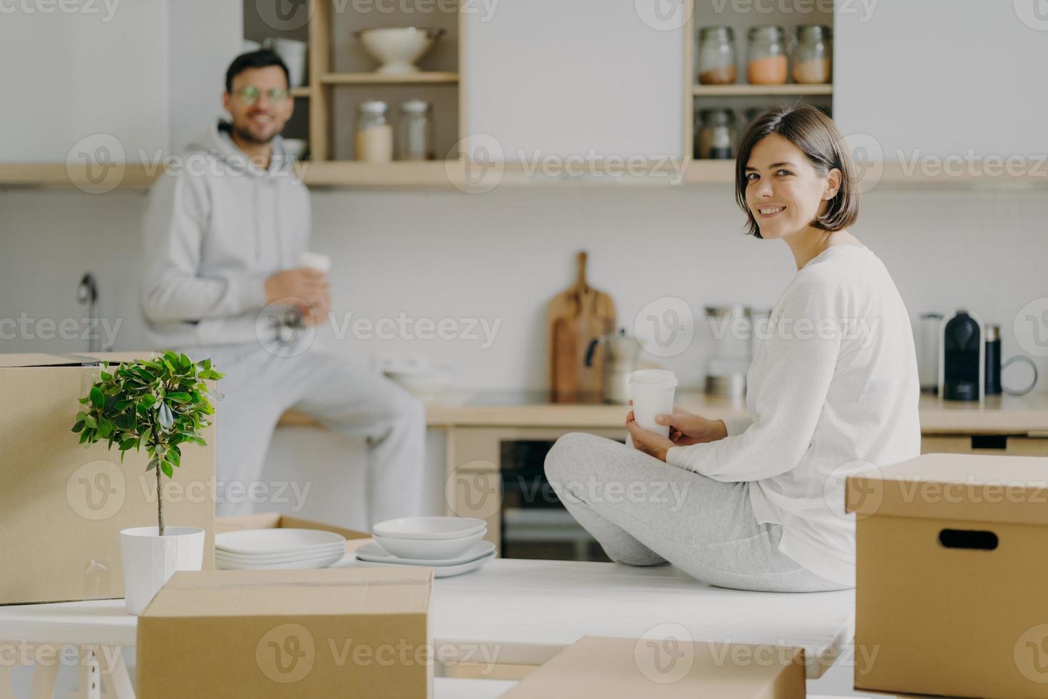 Horizontal shot of happy young woman and man sit on kitchen desk, drink coffee, have break after unpacking boxes, have pleasant talk, surrounded with plates, indoor plant in pot. Moving concept photo