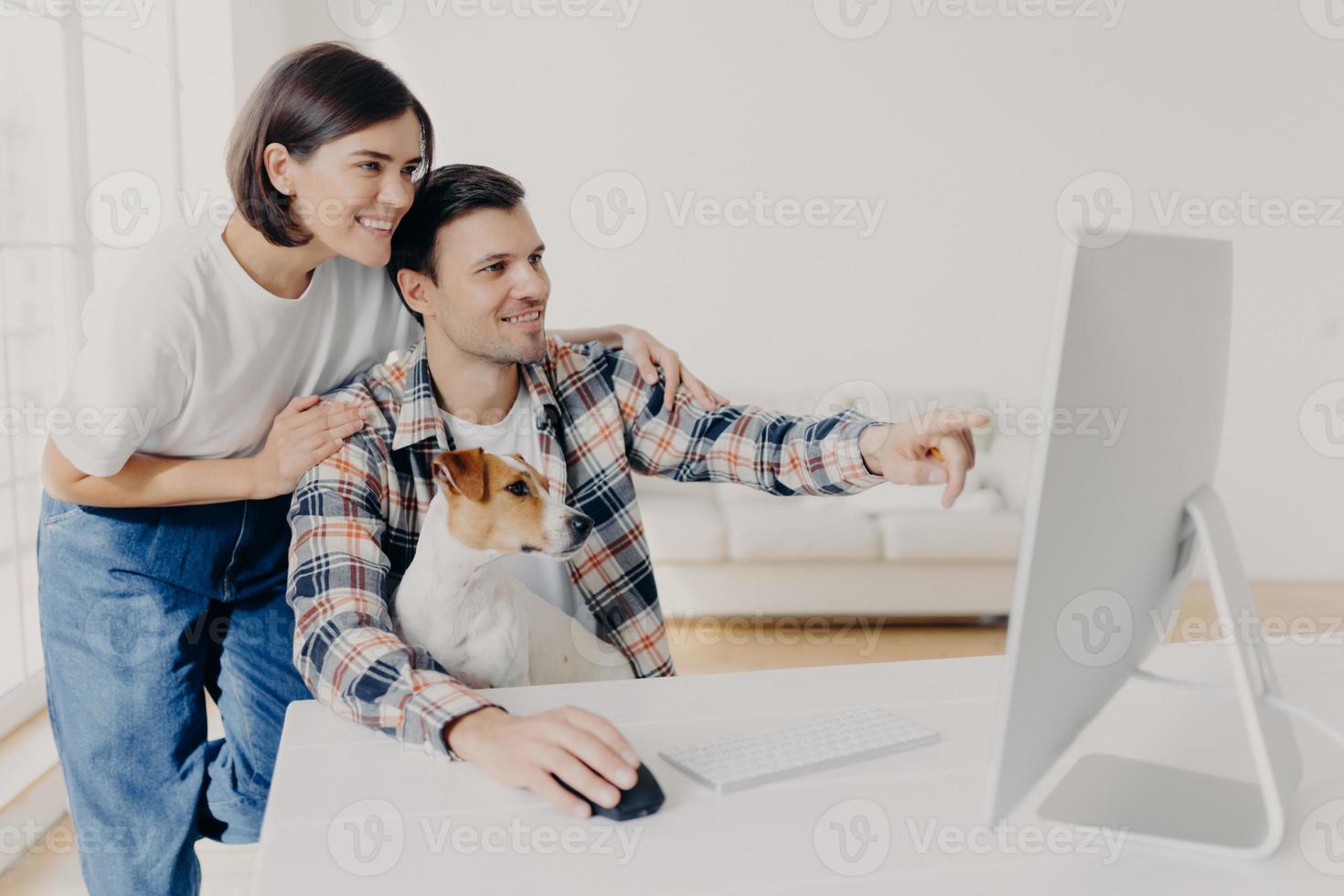 Happy man and his girlfriend consult about future project, point into monitor, pose at workplace together with dog in home interior, watch online tutorial video, spend time in modern apartment photo