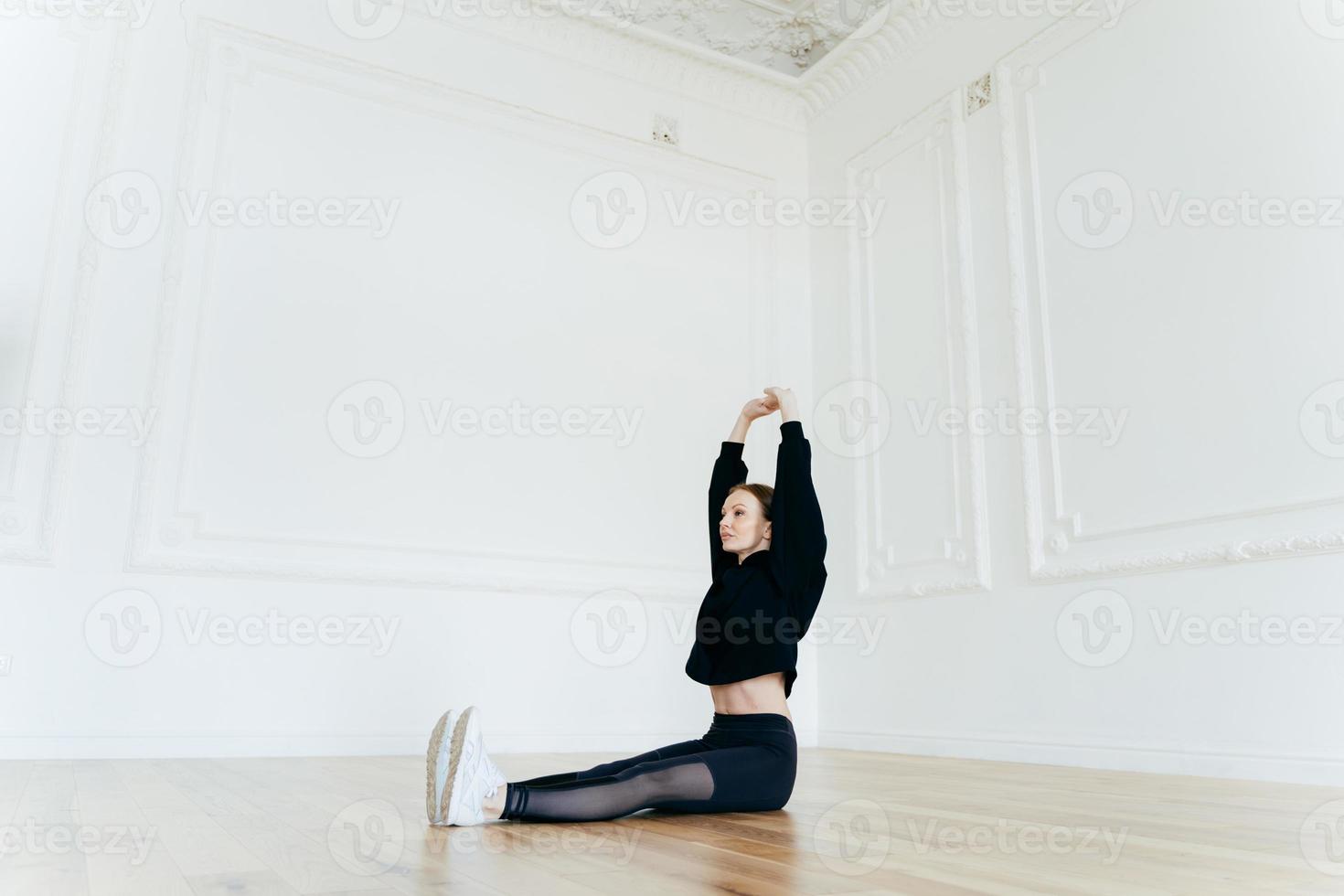 Young motivated fitness woman raises hands, does stretching exercises on floor, wears black sweatshirt, leggings and sneakers, has slim body shape, sits in spacious hall. Sportsgirl trains indoor photo