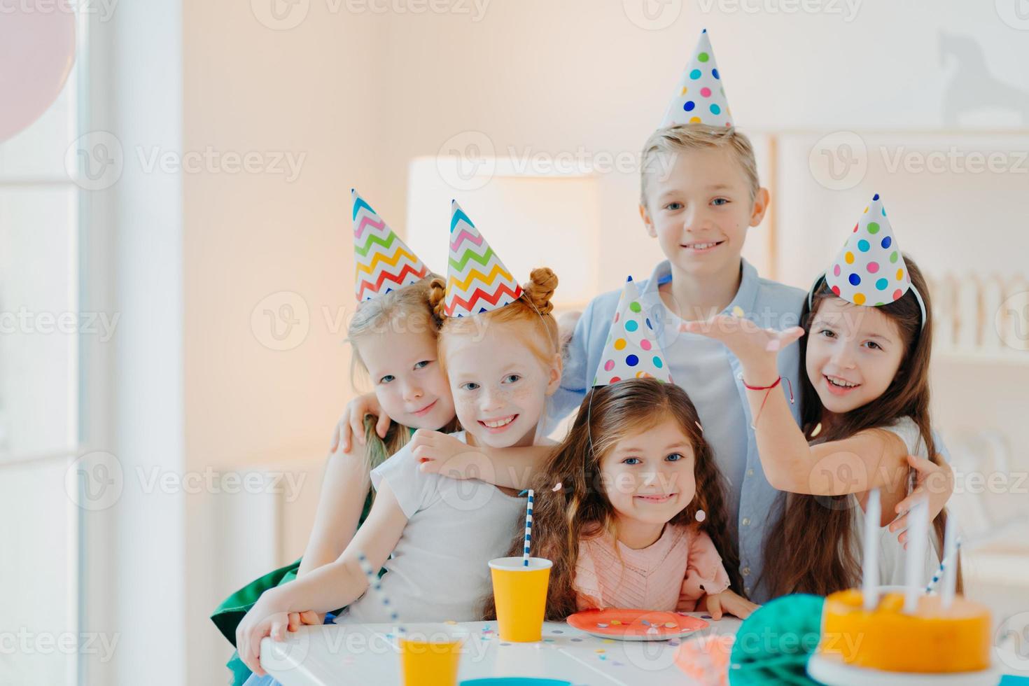 Indoor shot of happy kids celebrate party with falling confetti, wear cone party hats, pose near festive table with cakes, embrace and pose together. Childrens birthday photo