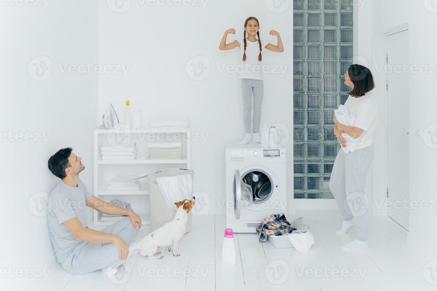 Photo of happy schoolgirl poses on top of washing machine, shows muscles, raises arms, ready to help parents with washing or laundry. Woman and man pose in washing room with dog and small kid