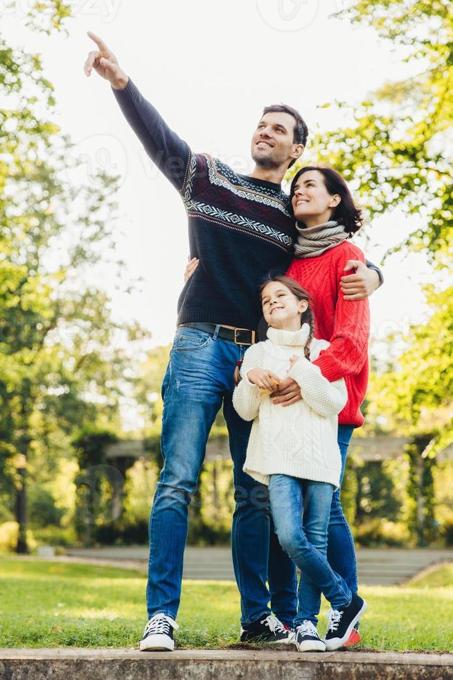 Vertical portrait of family of three members stand close to each other, embrace, have walk across autumn park, notice squirrels on tree. Friendly father, mother and daughter have good relationship photo