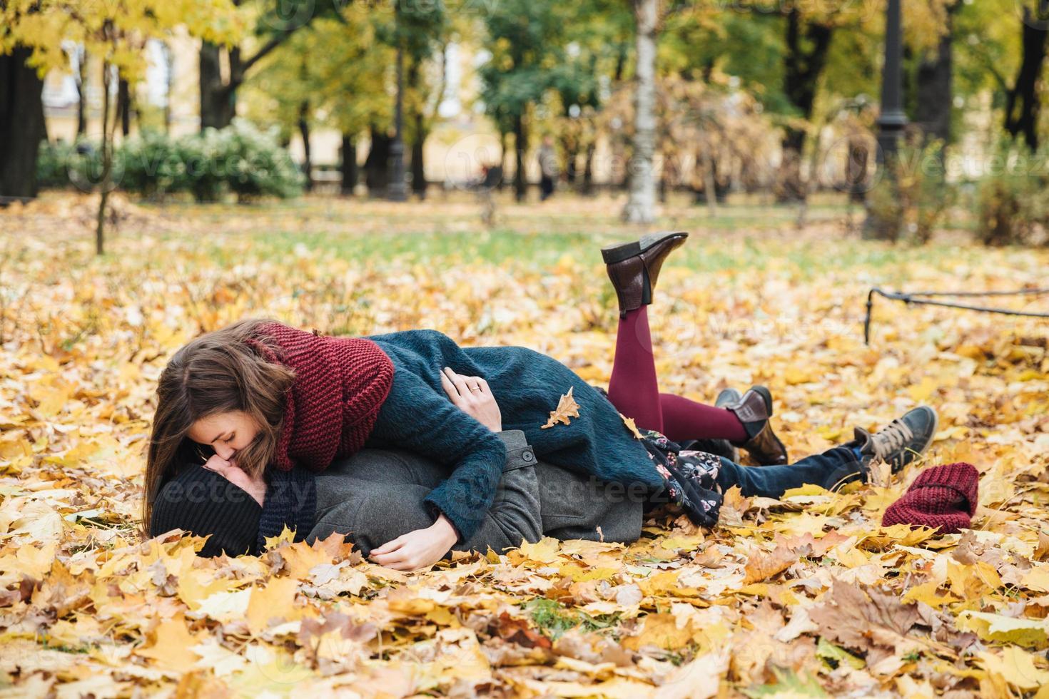 Boyfriend and girlfriend have romantic kiss as lie on yellow leaves in park, spend sunny autumn weekends together, have good mood. Couple in love enjoy togetherness outdoors. Season and people photo