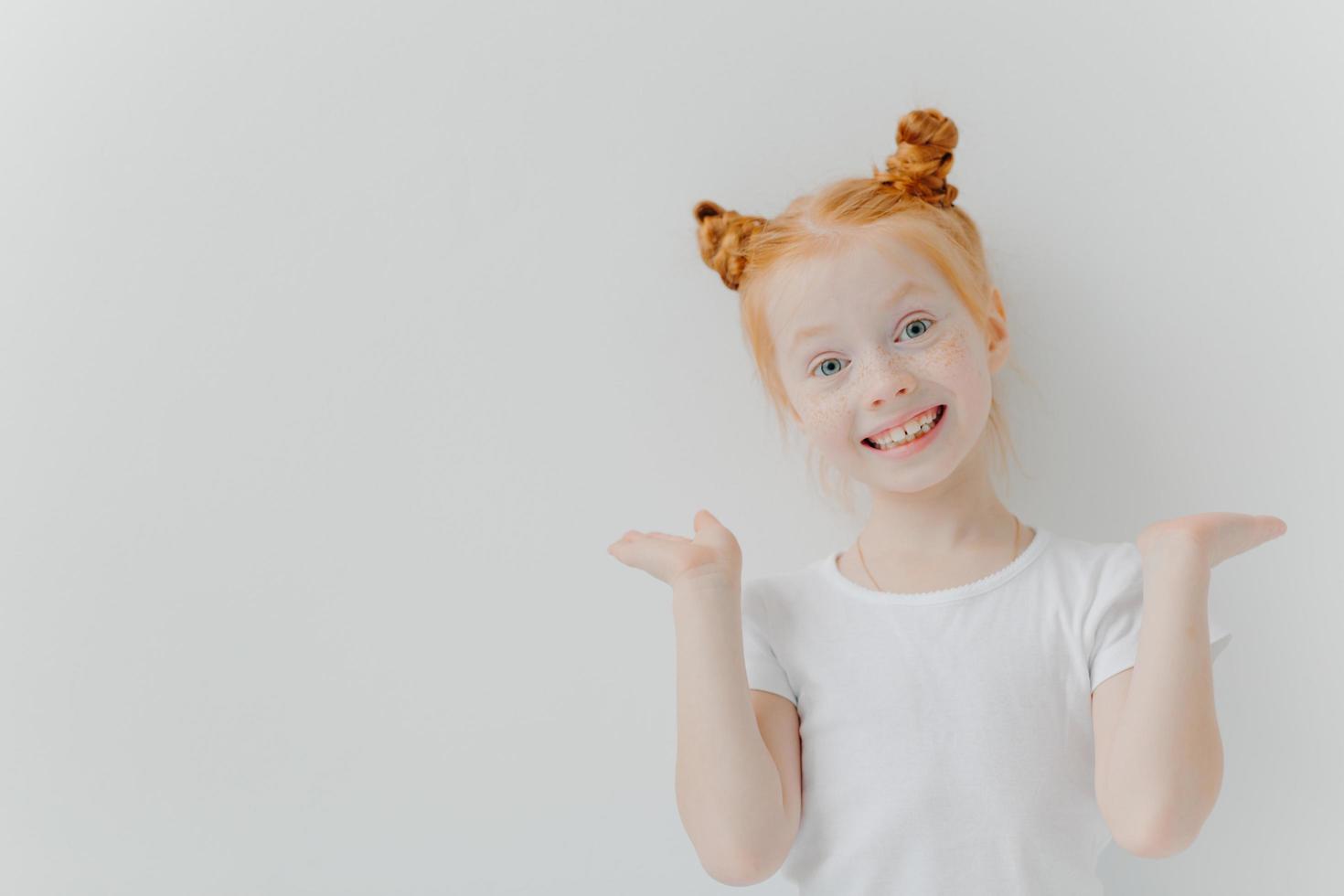 Playful red haired girl raises palms, has double buns, looks happily at camera, wears casual white t shirt, poses indoor, makes funny face, has toothy smile, freckled skin. Children, emotions concept photo