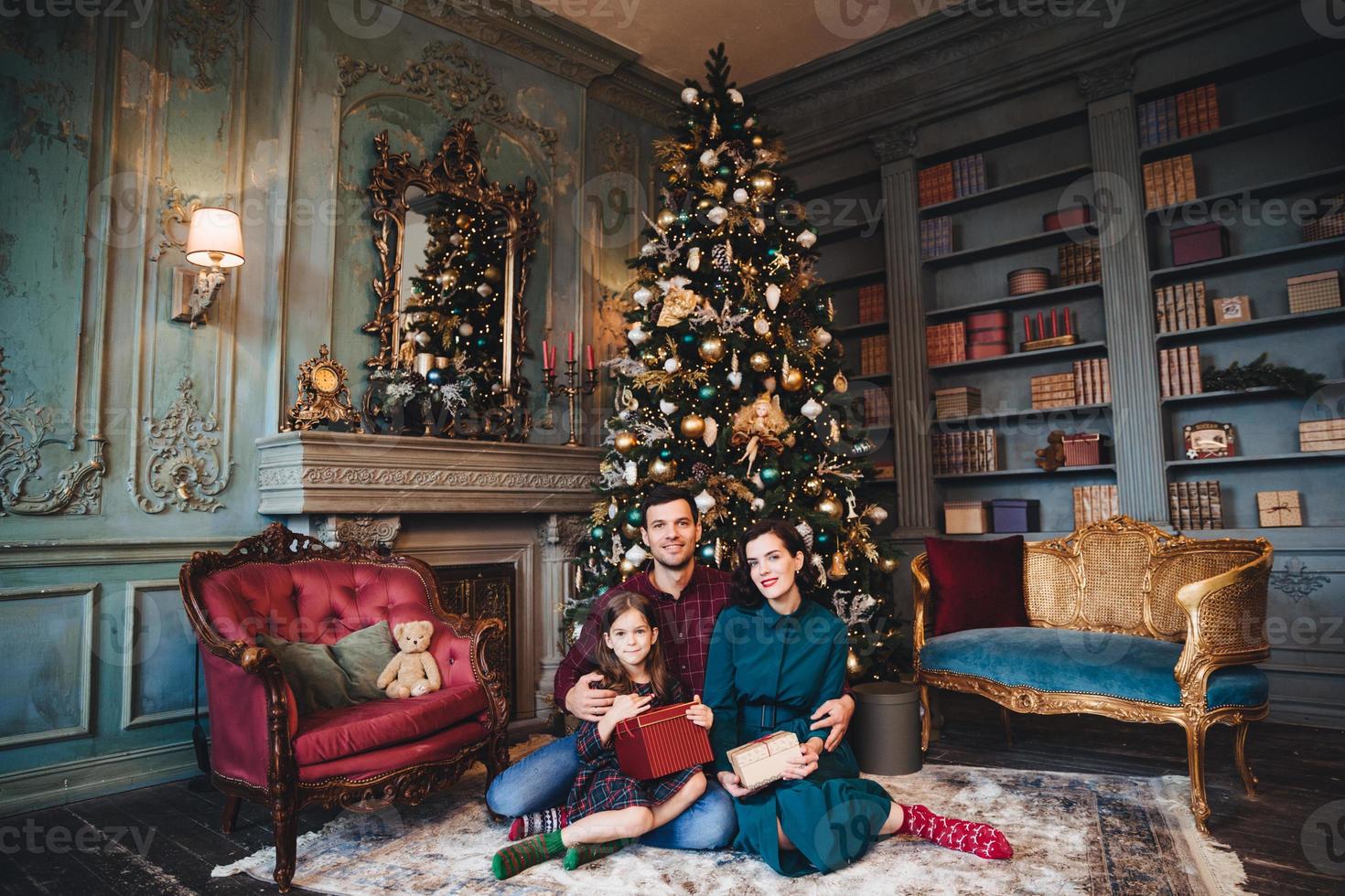 Friendly family sits near decorated Christams tree, embrace each other, hold packed presents, enjoy majestic atmospere. Father, mother and daughter near New Year tree in living room. Festive concept photo