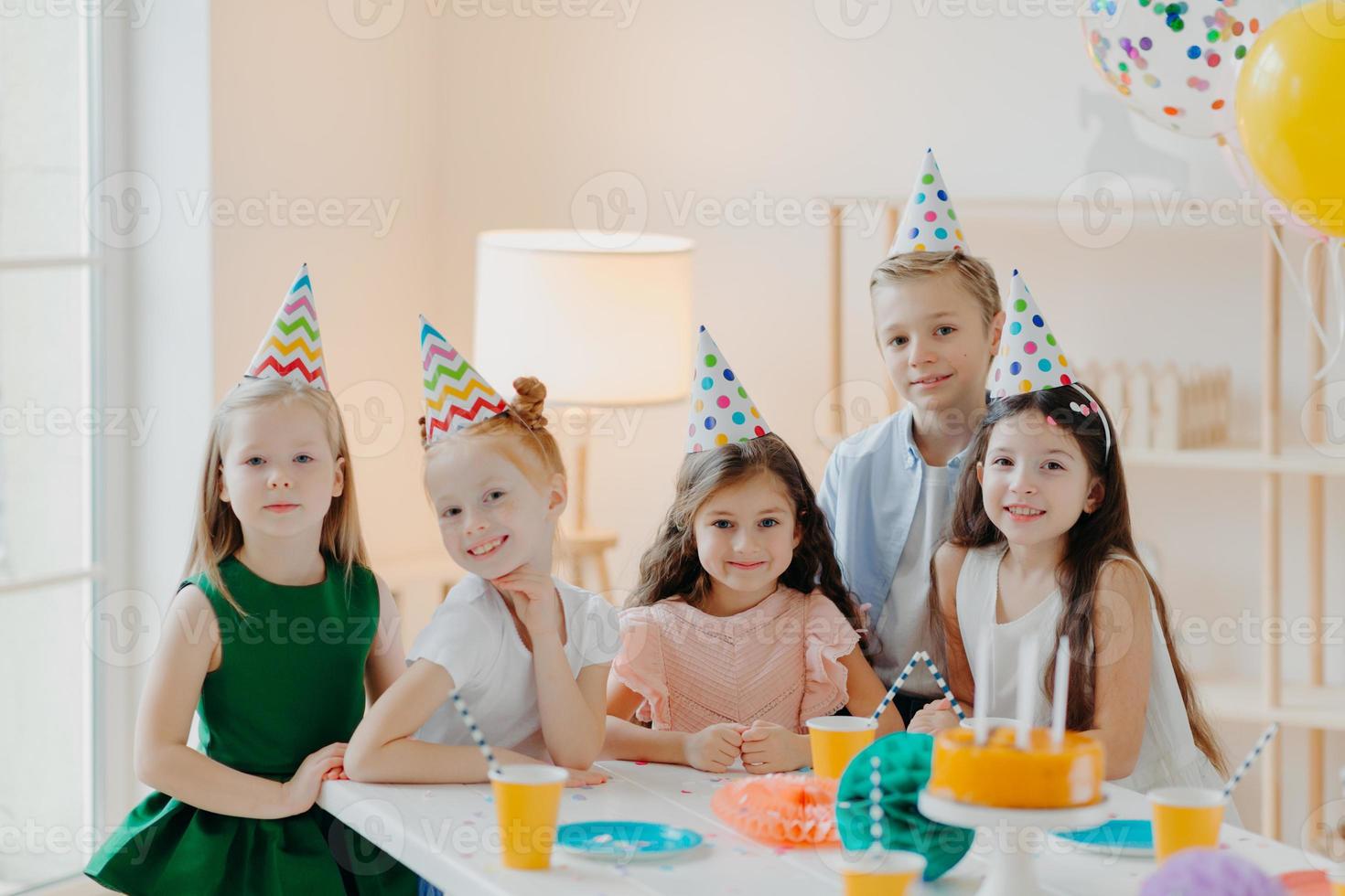 Children, celebration and birthday concept. Positive children have fun together at party, wear cone hats, eat delicious cake, play interesting games, being in good mood photo