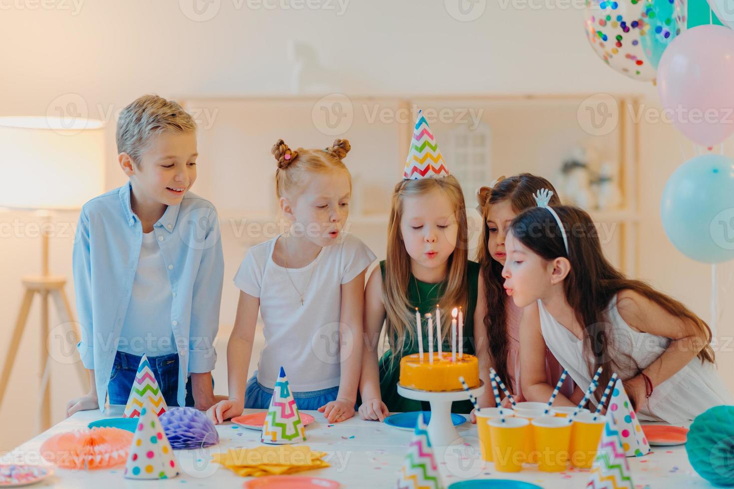 los niños pequeños celebran la fiesta de cumpleaños, soplan velas en el pastel, se reúnen en la mesa festiva, tienen buen humor, disfrutan pasar tiempo juntos, piden deseos, usan sombreros de fiesta, posan en el interior con globos inflados foto