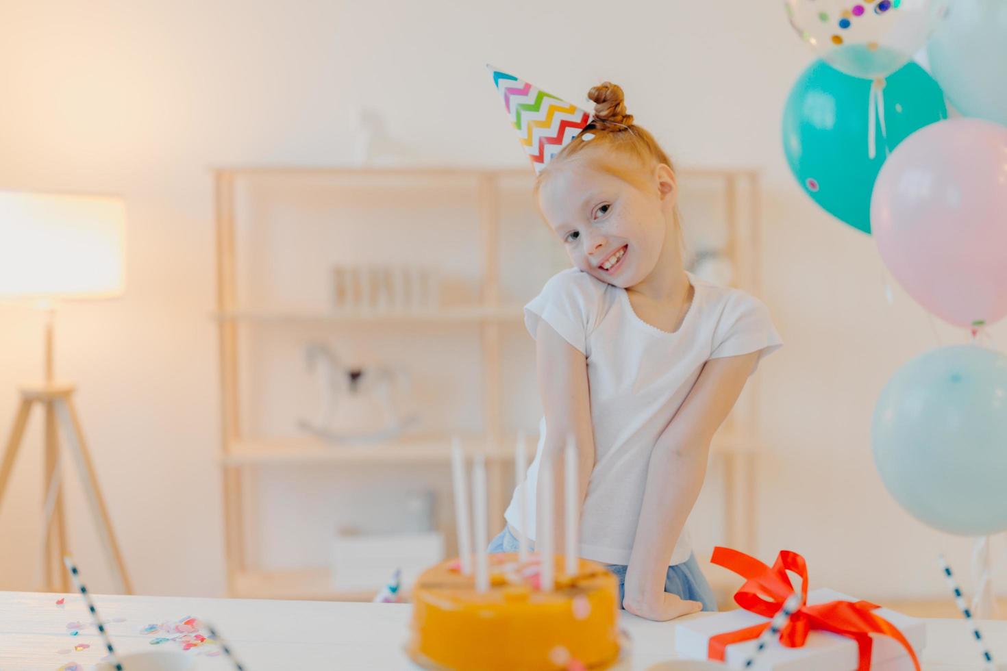 Little ginger girl wears party hat and white tshirt, stands near festive table with cake, blows candles and makes wish during her birthday, poses in white room with inflated balloons, smiles joyfully photo
