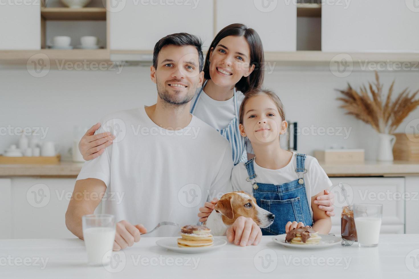 Family, leisure, pastime, eating concept. Father, mother and daughter, jack russell terrier dog pose all together at camera against kitchen interior, get pleasure from eating pancakes drink fresh milk photo