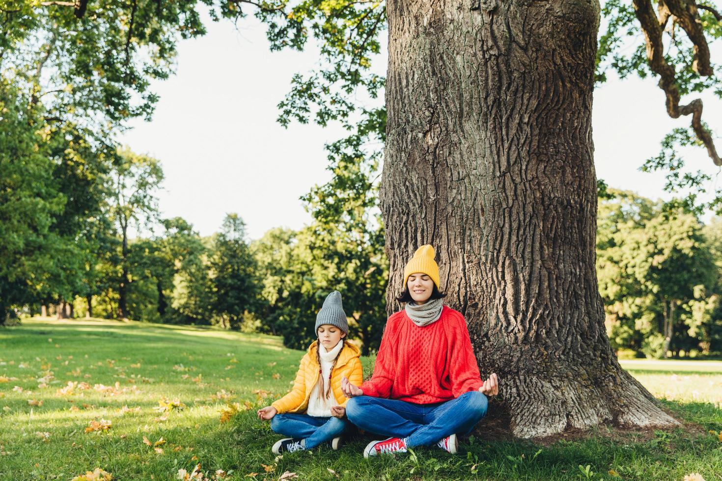 Yoga and meditation concept. Beautiful young woman in knitted clothes and small child keep eyes closed, meditate outdoors in green park, feel relaxed, calm and peaceful, keep hands in mudra sign photo