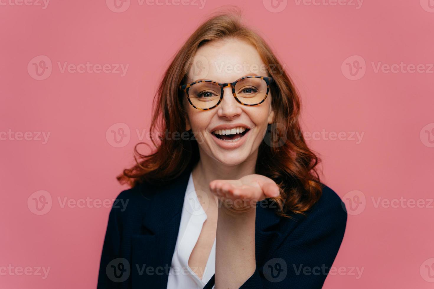 Photo of red haired beautiful woman stretches palm over mouth, sends air kiss to beloved person, has glad facial expression, ginger curly hair, wears formal clothes, isolated on pink background