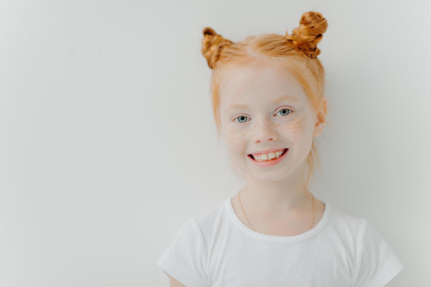 Positive attractive little girl with double ginger buns, freckles on face, toothy smile, dressed in casual t shirt, stands against white background, empty space for your advert. Children concept photo