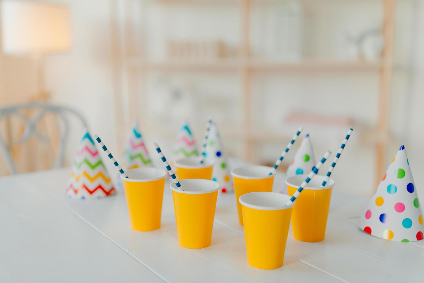 Holiday hats and paper cups with beverage and straws on white table. Festive event. Birthday party celebration concept. Blurred background. Nobody on picture photo