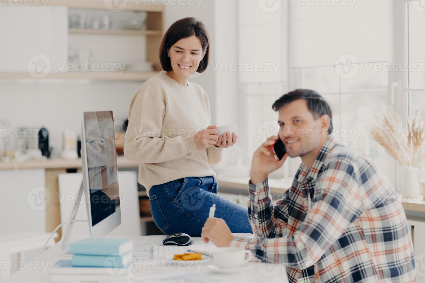 Horizontal shot of cheerful man has telephone conversation, discusses financial contract agreement, dressed in checkered shirt, writes down information, happy woman holds mug of drink, leans at table photo