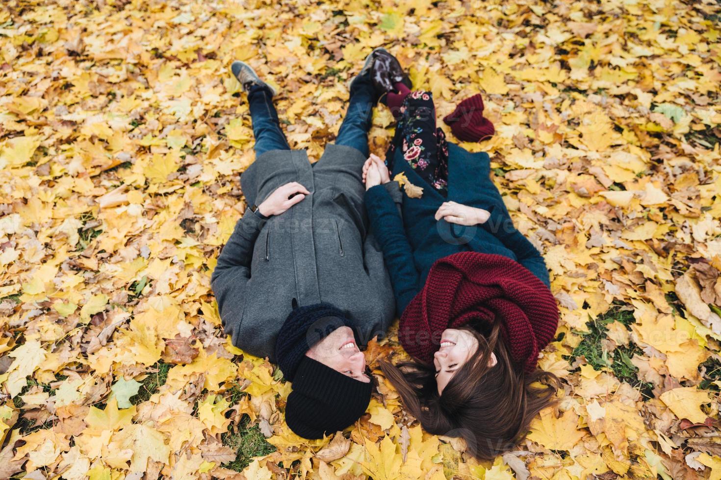 una pareja joven y romántica se acuesta en hojas amarillas en el parque, se toman de la mano, se miran con gran amor, disfrutan del día soleado de otoño. concepto de personas, relaciones, unión y romance. foto